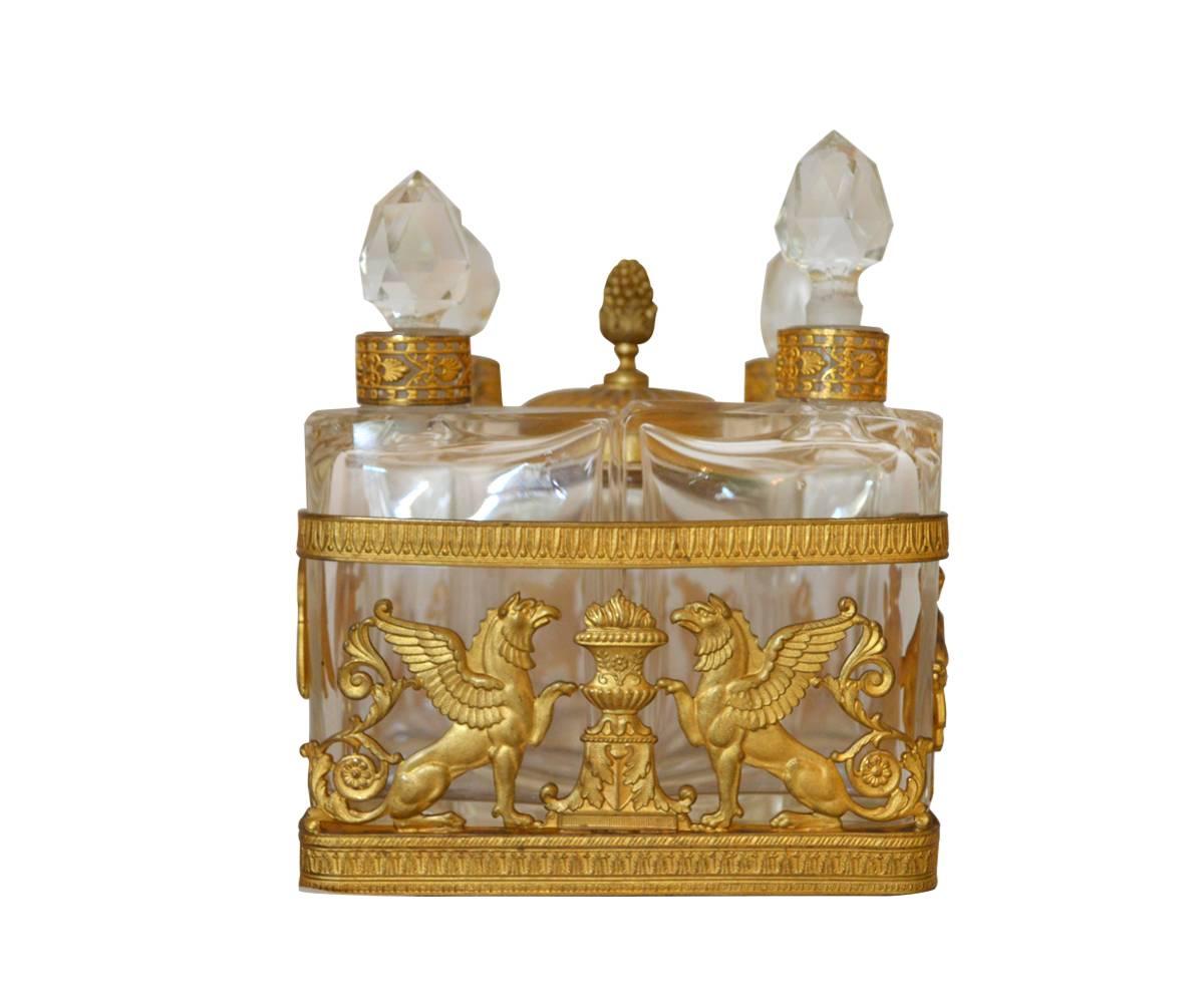 This empire style perfume bottle caddy has four perfume bottles and a space in the middle to hold cotton balls. It is decorated with griffins on one side. Another side has a lady reclined with a mirror in her hand and an angel brings her a bowl vase