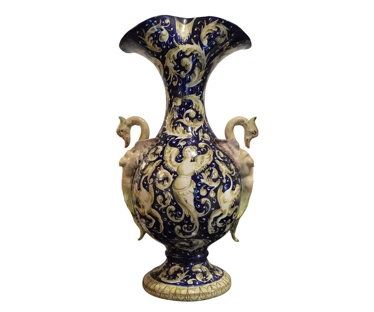 This magnificent Majolica vase features gorgeous dark blues in the background with green and ivory accents. The griffin handles adorn the sides. The back features a winged angel and more griffins and the front painting features two happy maidens in