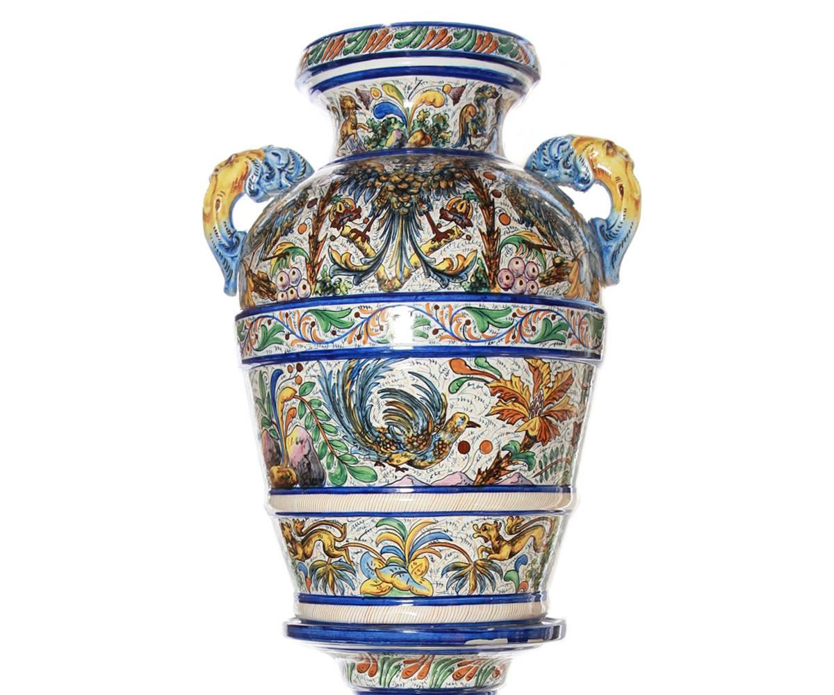 This large pair of Italian hand-painted ceramic urns in the Majolica style feature faces as handles. The brightly hand-painted images include birds, lions and various foliage. The colors include vibrant blues, yells and greens.