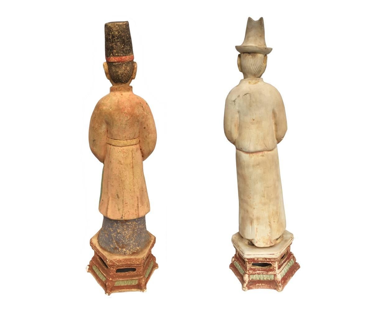 Pair of tall Ming dynasty pottery figures; each standing on hexagonal bases, wearing long robes and cap and each holding a gift or offering piece, some remains of original painted decoration, heads typical removable.