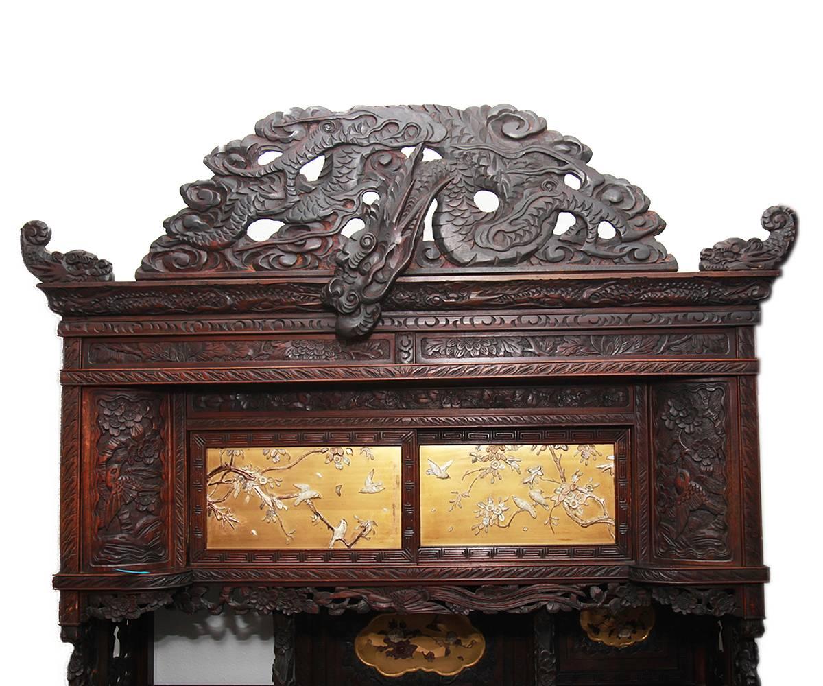 Antique Japanese, very elaborately carved display cabinet; with carved dragon crest and relief bird and foliage motifs: Six doors with plaques having Shibayama_ style carved applique bird and flower motifs on gold lacquered grounds, various shelves(