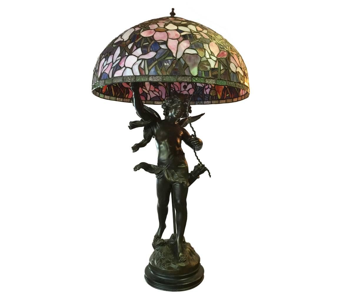 Offered is this magnificent antique bronze sculpture which has been professionally converted to a lamp featuring a cherub with archery accents. This winged angel has delightful character and rests on a bronze base. The stained glass shade appears to