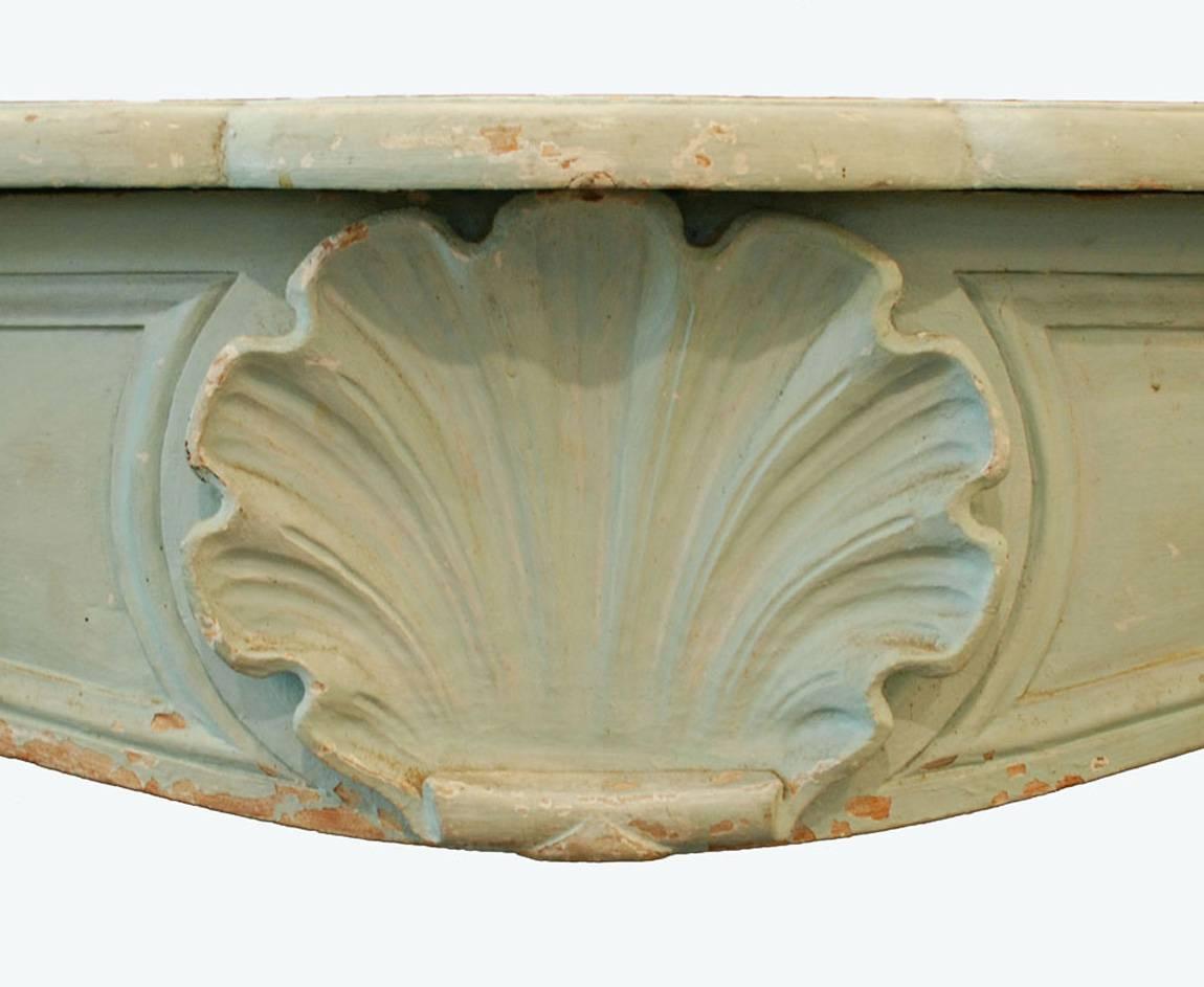 An understated antique wooden fireplace surround in mint. The frieze is decorated by a single scallop shell in relief and minimal carving to emphasize the lines of the piece. This carving appears across the full front of the mantel, as well as a