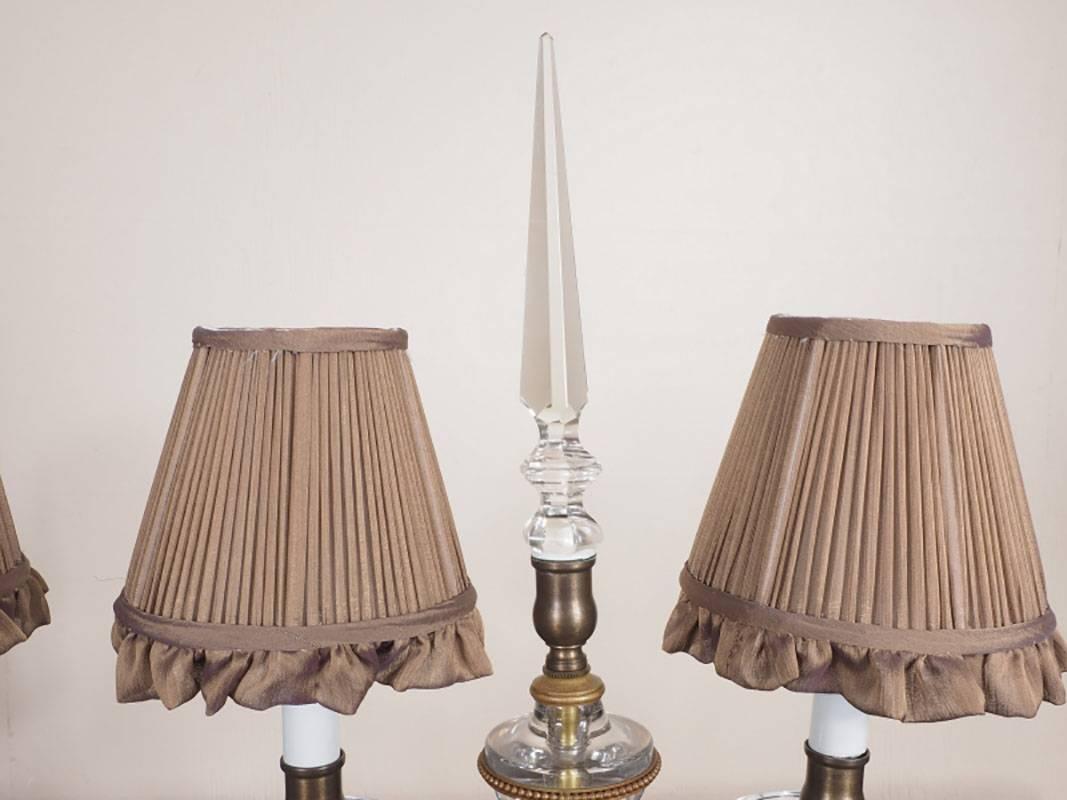 Crystal lusters (one missing) hang from the drip cups on these two-arm candelabra shaped glass and bronze lamps. Large spire finial stands out of the urn-shaped crystal center with bronze beaded banding. Custom gathered silk lamp shades are included.