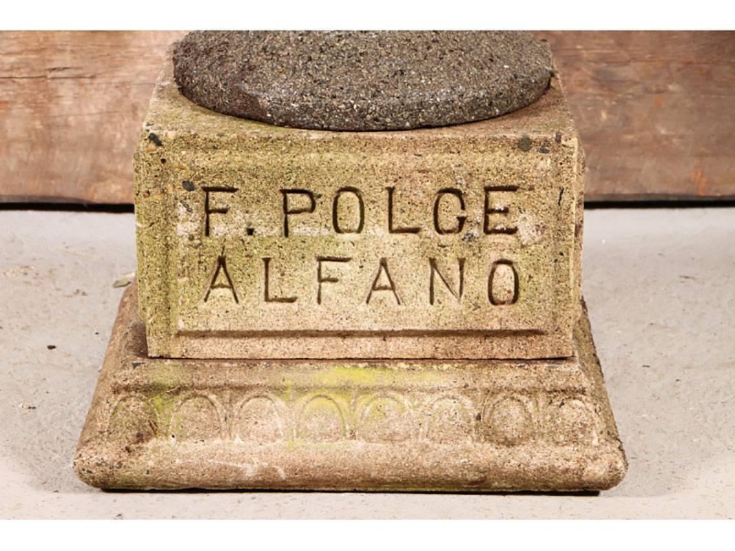 A stone and cement urn with grape leaves, neoclassical lines and molded handles sitting on two-part carved stone short pedestal. The inscription reads, 