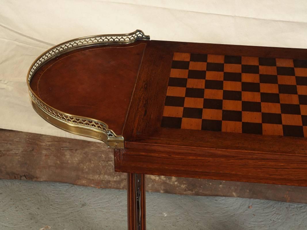 Regency style games table having an ovoid form with leather covered side panels with brass gallery, string inlay and supported by wood brackets. The surface with an inlaid game board. The game table is supported by two string inlaid decorated