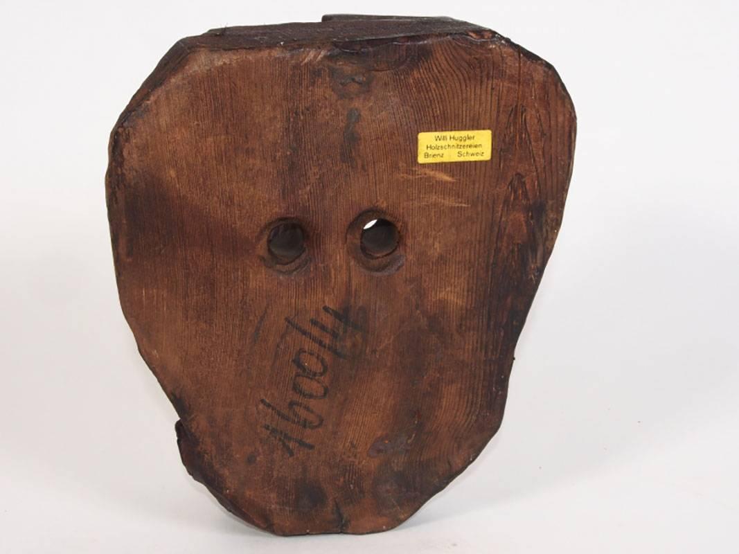 Carved grotesque wood mask with pierced eyes, very hooked nose, cigar between very crooked bone teeth. Label on verso reads, “Willi Huggler (Swiss, 1925-2016), Holzschnitzereien, Brienz, Schweiz