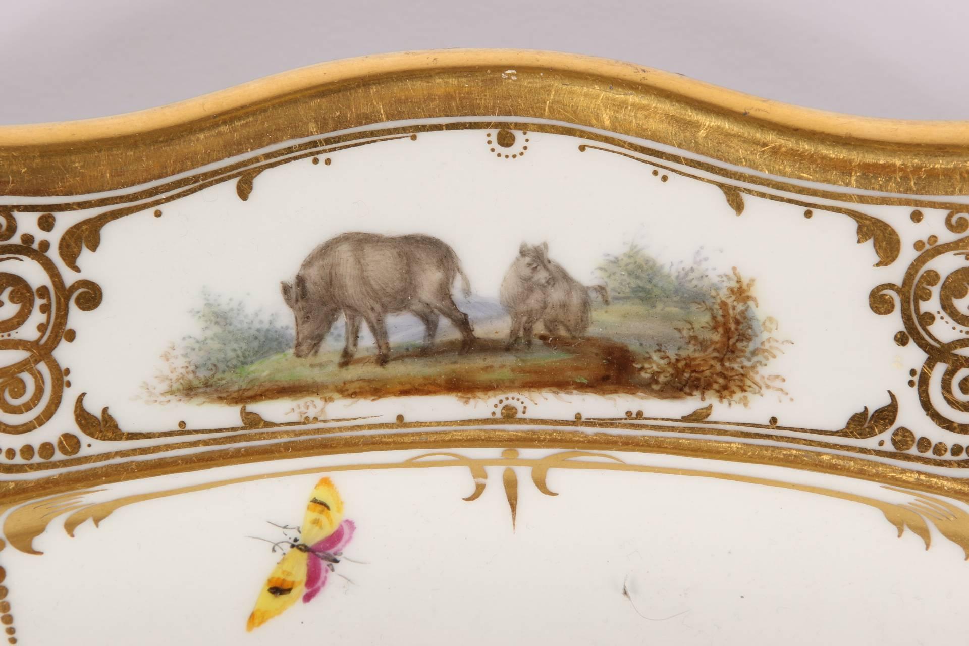 Probably custom-made oval form with gilt scalloped rim, the central scene depicting three boars in a forest clearing. The broad rim with four cartouches illustrating pairs of boars including babies, and one with a dog approaching a boar. Gilt