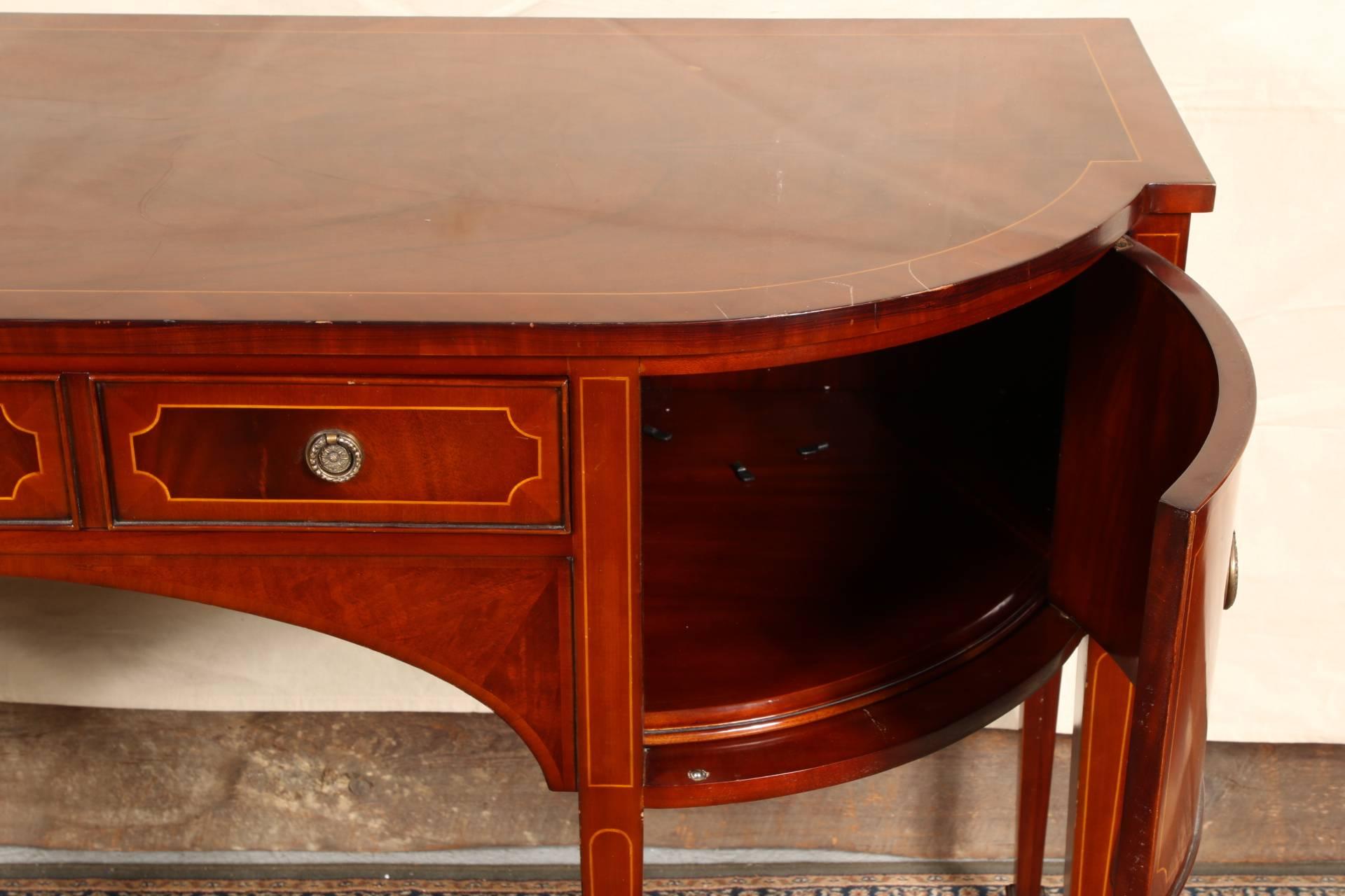 Features tonal mahogany veneer with string inlay accents, a central tri-panelled lined front drawer, two side cabinets, tapered legs and brass ring pull hardware.
Exhibits surface scratches and losses to the veneer.