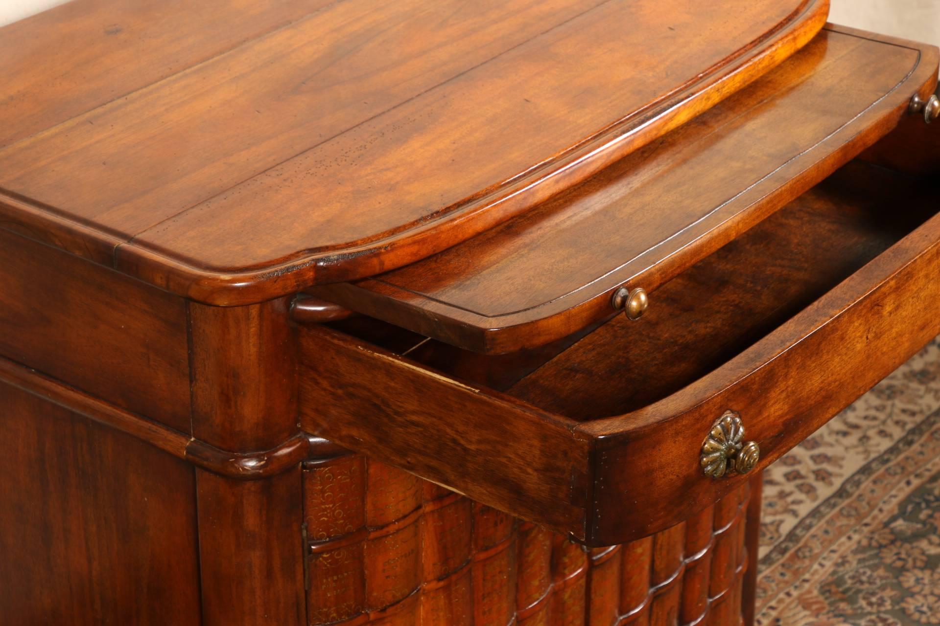 A shaped top with a slide pull and a single drawer over a cabinet base. The double doors with carved and gilt faux book bindings. Raised on turned legs in front and square ones at back, metal knob pulls. Paper label on the bottom of the