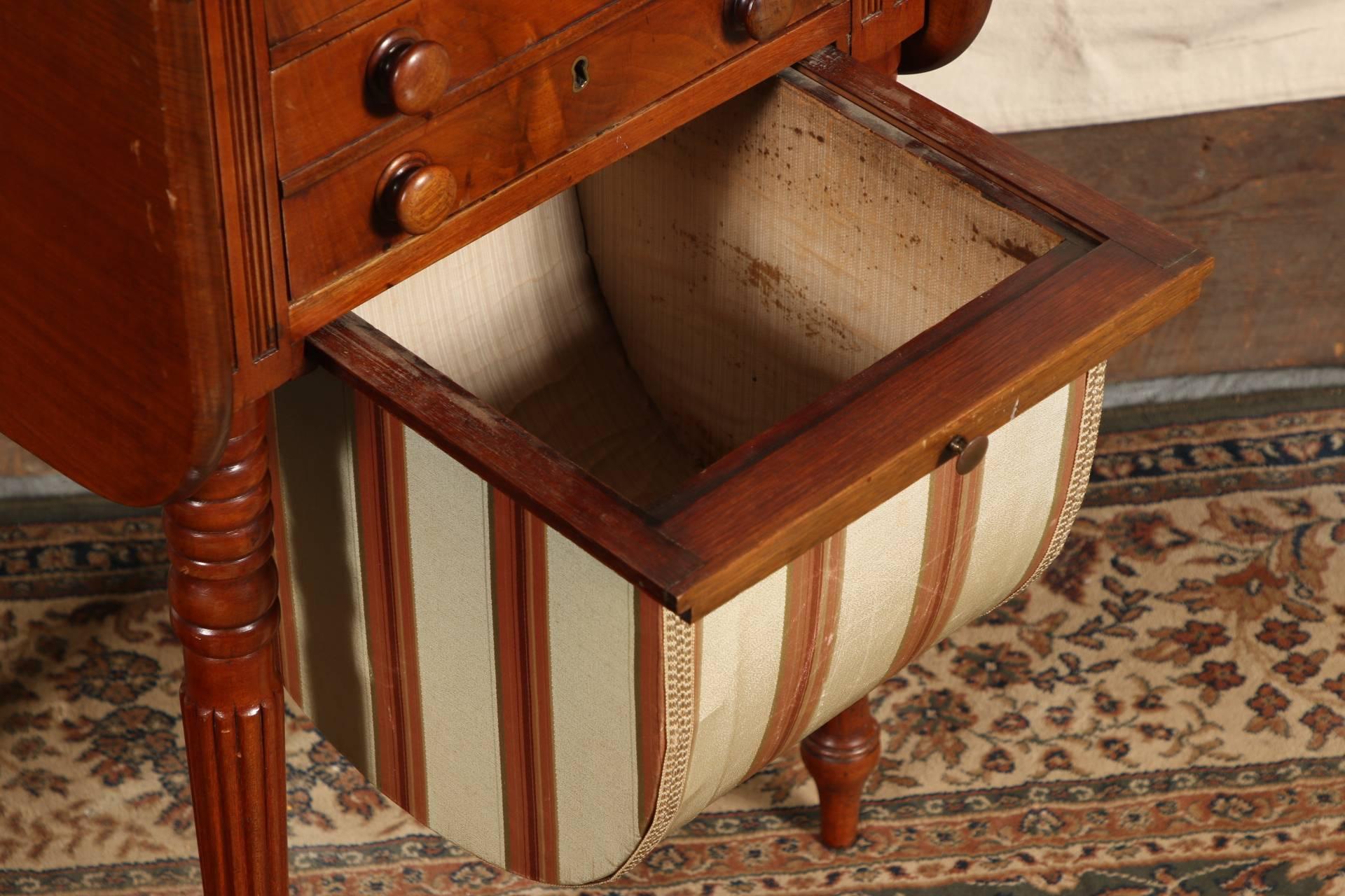 Mahogany with drop leaf sides, the cabinet with reeded supports over turned and reeded cylindrical legs with peg feet. Three drawers with turned pulls and brass keyholes over a pull-out lower striped fabric compartment.
Condition: joinery loose