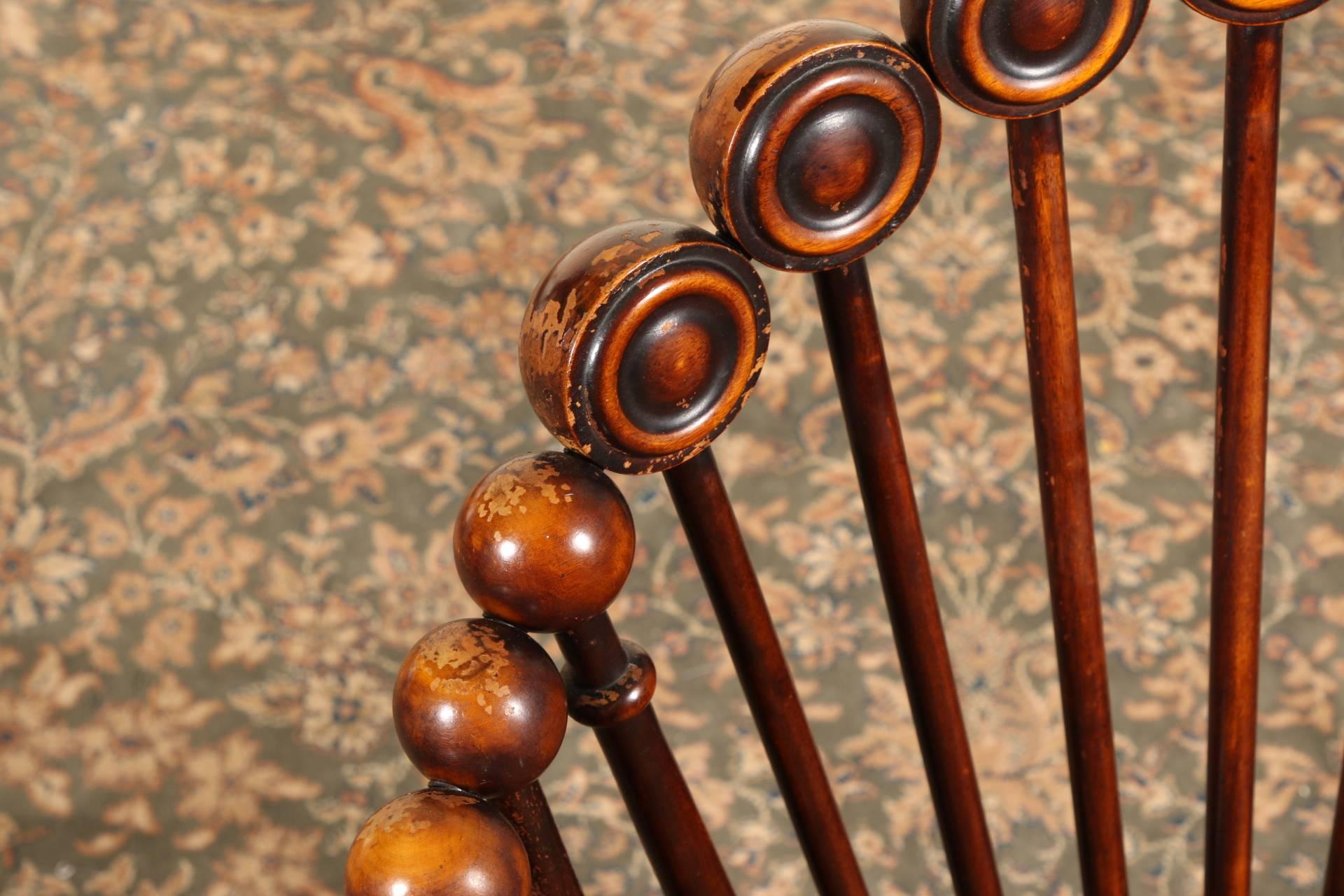 Dark stained and turned maple tub back shapes. The backs with flat half spheres, the sides and bottom rungs with ball forms, all on sticks. Turned arms and raised on shaped front legs on casters, the back legs square and splayed. Peach tone