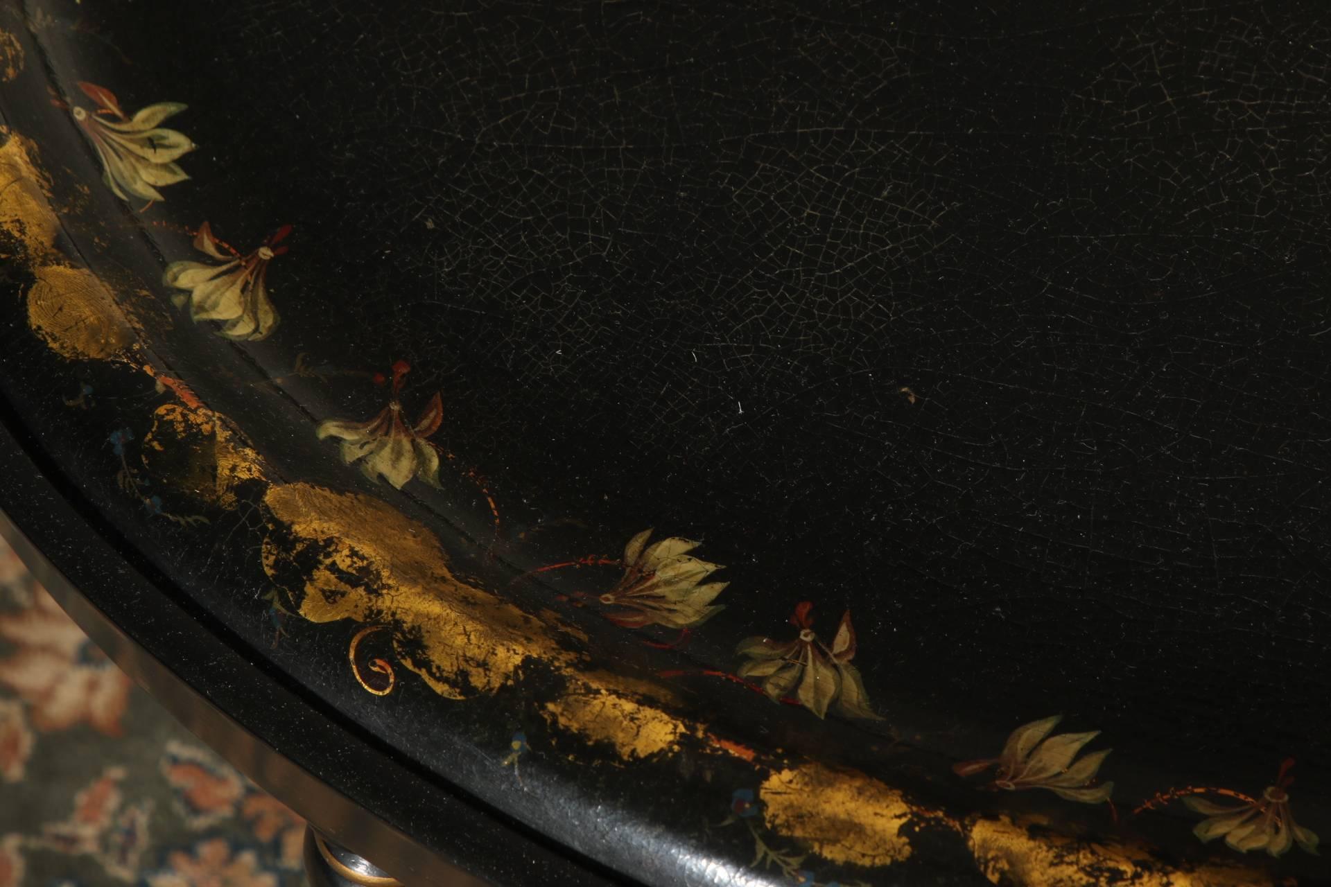 Ebonized oval with the tray inset in one piece with the stand. The tray with raised border decorated with a band of clusters of gilt leaves with pendant red and white flowers. The ebonized stand with turned splayed legs with gilt details and faux