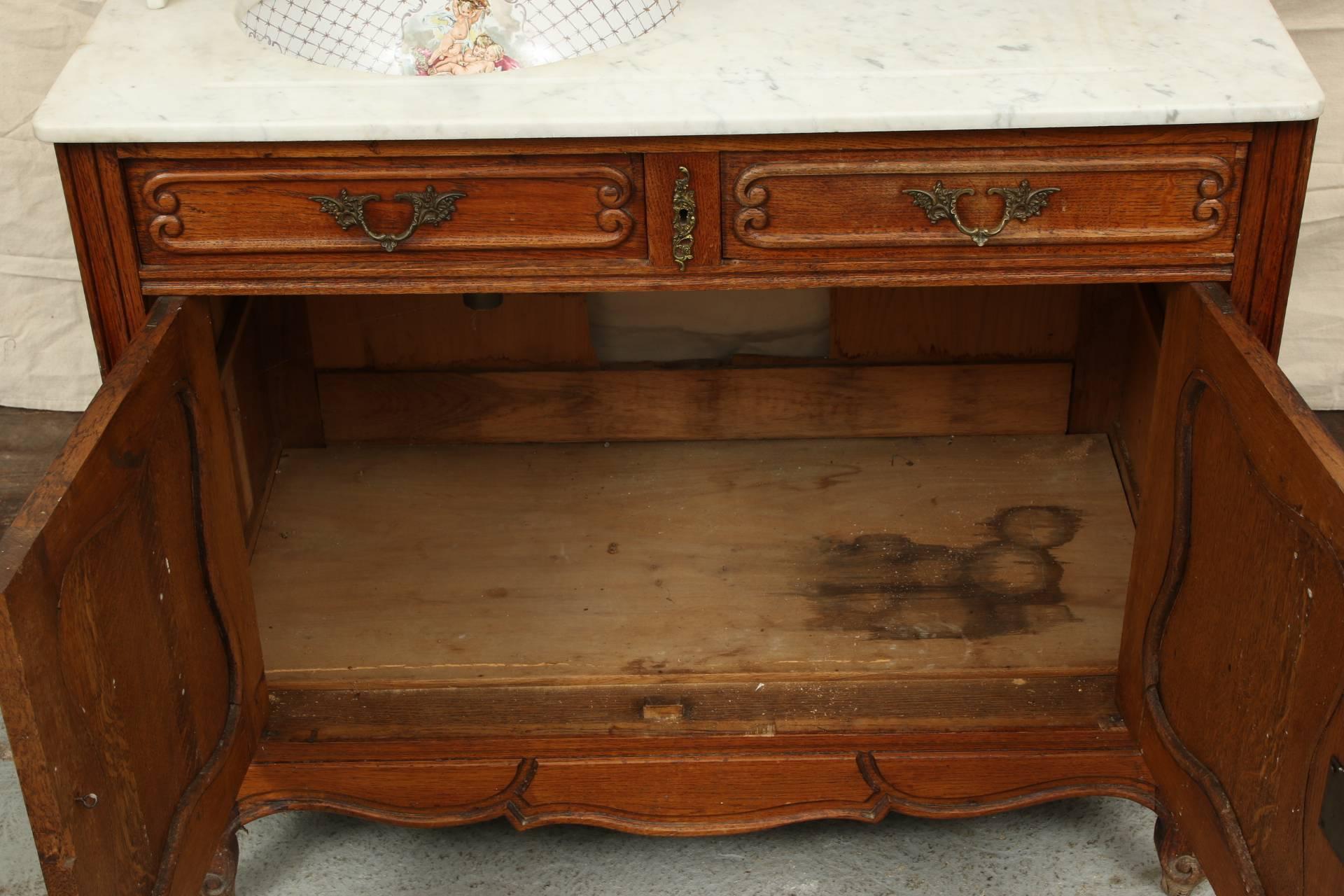 French country style double door oak cabinet with shaped framed panels and two short apron drawers. A triple hinged and bevelled mirror with carved frame and cartouche crest at the top back. A marble shelf with marble side supports sits above the