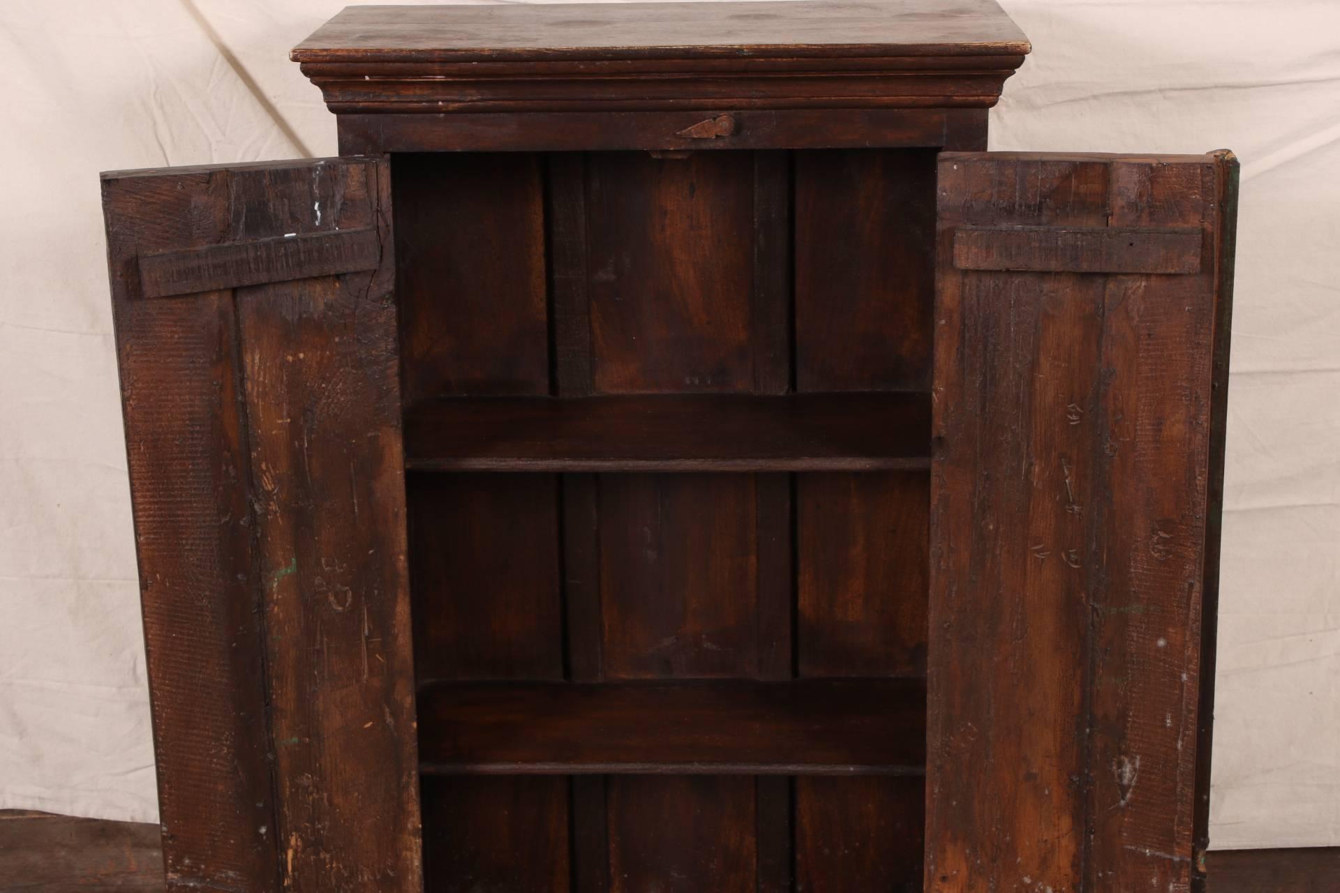 Dark stained wood with an overhanging top, carved double doors in green paint with tab closures above and below and iron ring pulls. Two shelves inside.
Condition: worn top, chip to right door top, wear to the base and weathered doors.