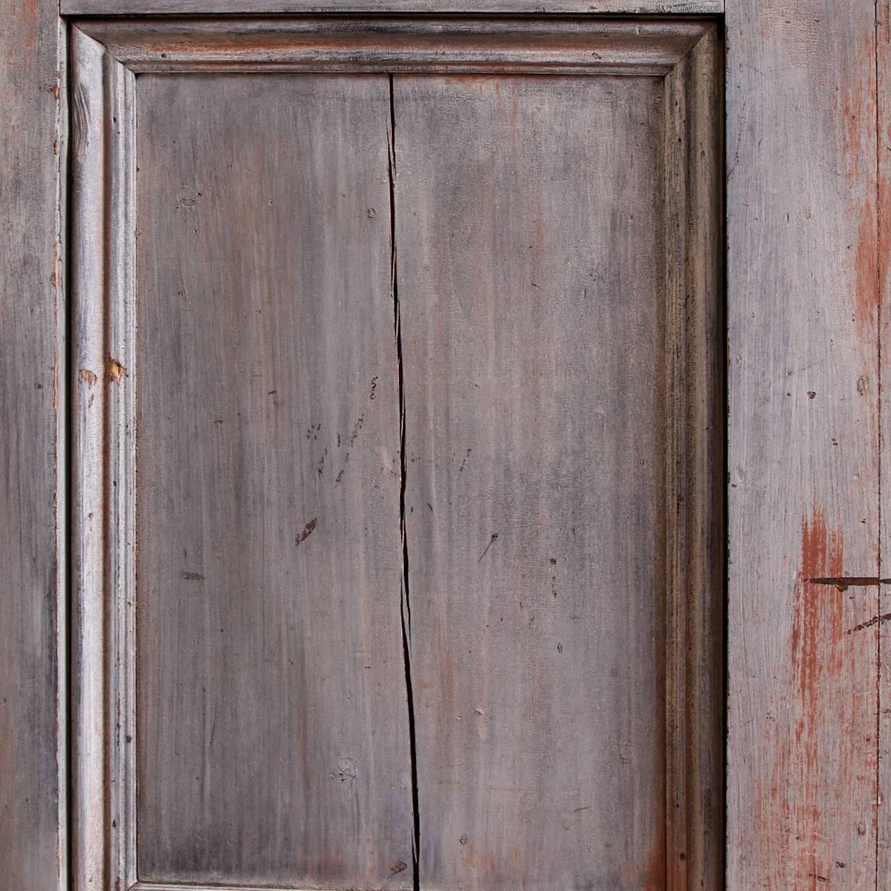 Two-part in a gray and pink weathered paint finish. The top part set back with a carved frame with openwork crest ornaments, two side sections with doors and a centre one with a glass paned door, all with escutcheons (with one key). The insides in
