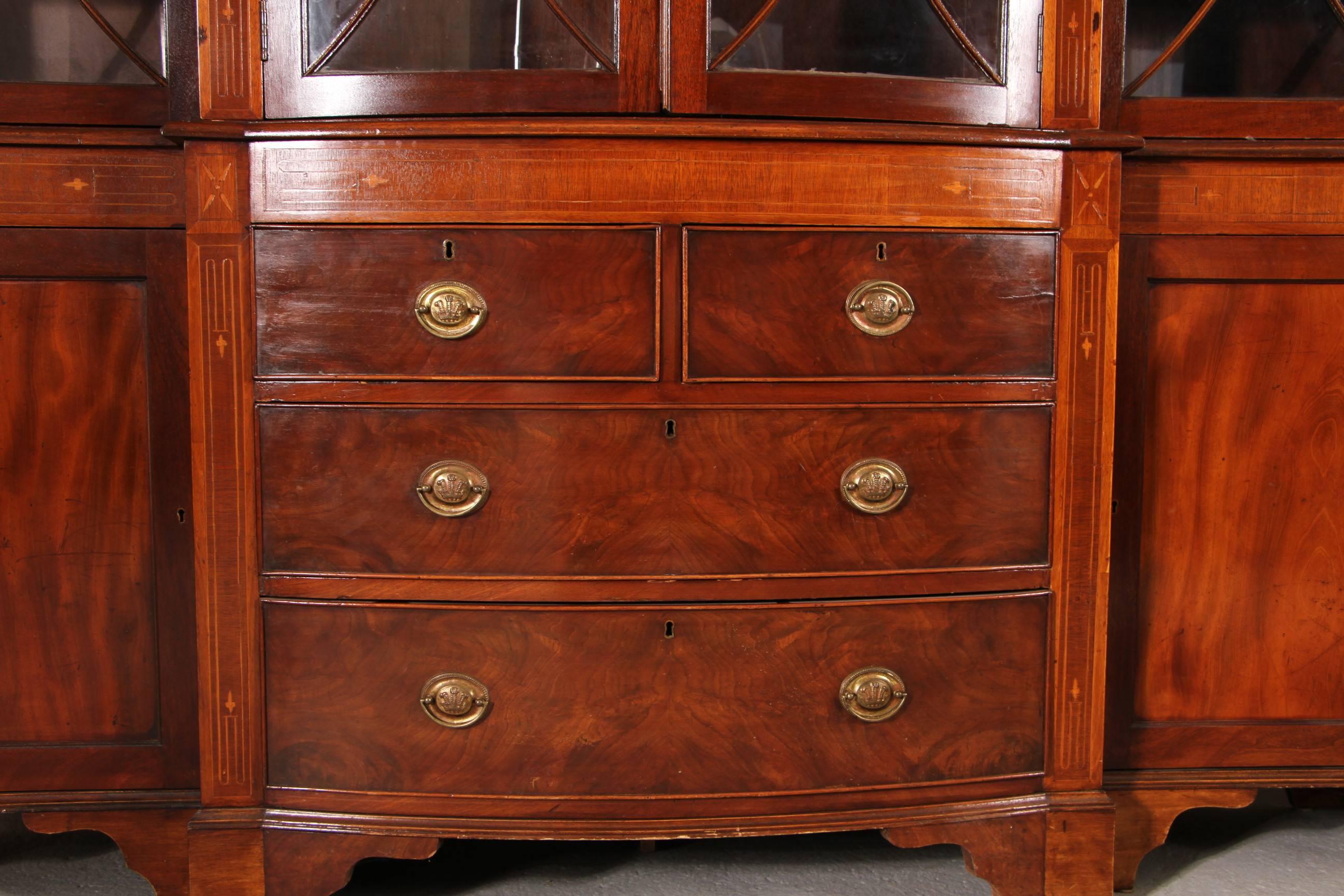 Beautifully finished and well proportioned antique mahogany English breakfront or bookcase with overall satinwood inlay, bowed center panels, divided glass paneled doors, scroll cut bracket feet, and decorative oval brass pulls. Age appropriate