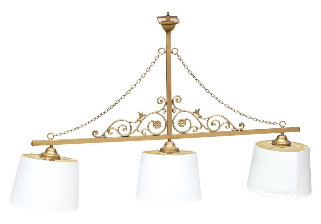 A three-light linear pendant chandelier, featuring a gold-tone metal frame with decorative chains, open scrollwork and three white hardback fabric shades. Designed for installation over a kitchen island or dining table, providing both functional
