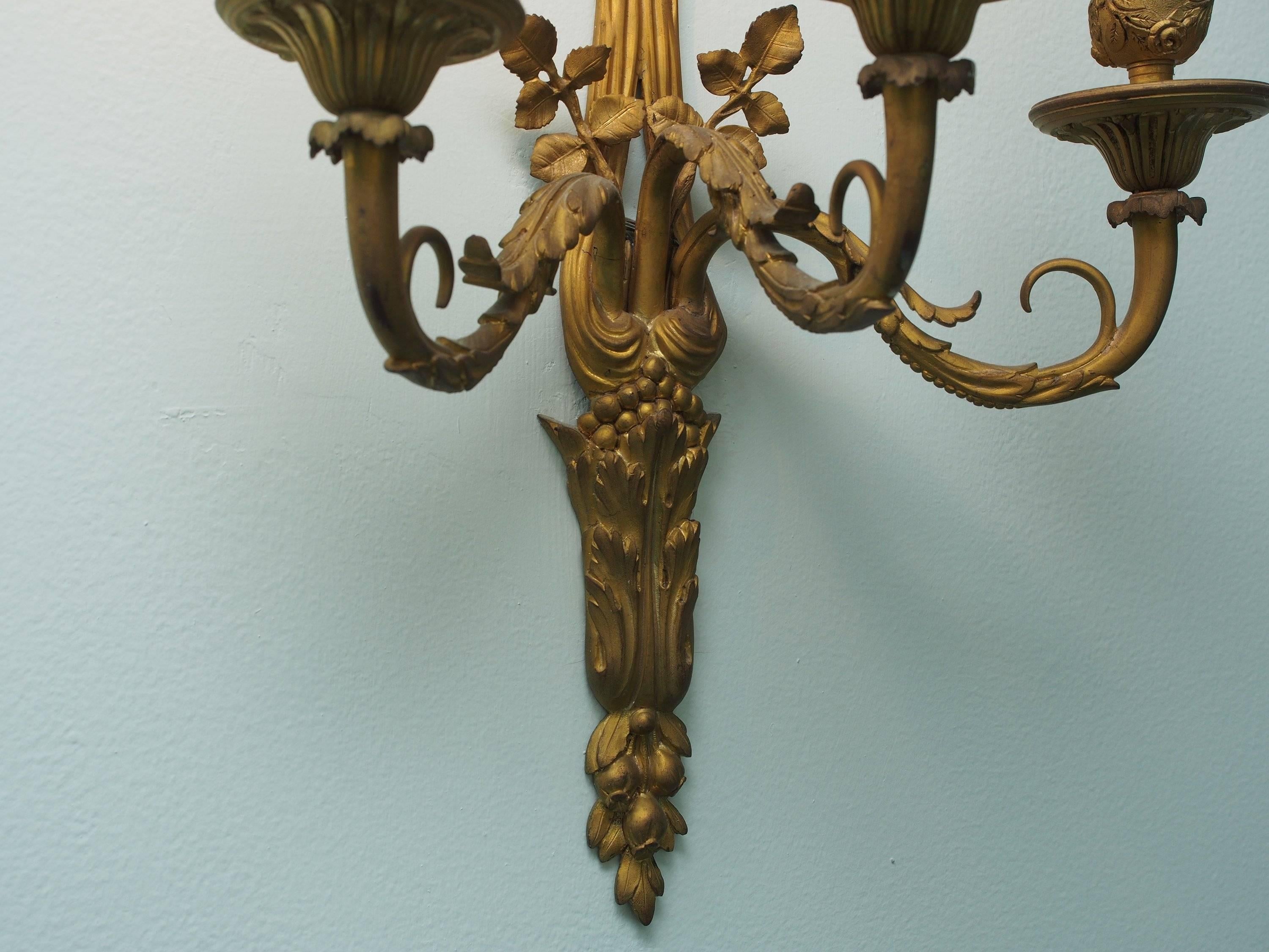 Pair of dore bronze wall sconces with tied ribbon crest, draped down and holding three arms, circa 1900-1920. The sconces with foliate lower column ending in three bell flowers, bobesche cast with roses, fluted caps and beaded trim. Each sconce