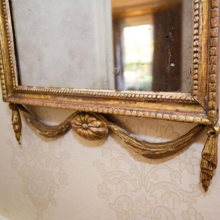 This antique Adam style rectangle carved giltwood frame mirror is headed by an anthemion spray atop an acanthus leaf spray. The molded frame is decorated with beads and is flanked by garlands of bell-flowers that continue down the sides. Drapery and