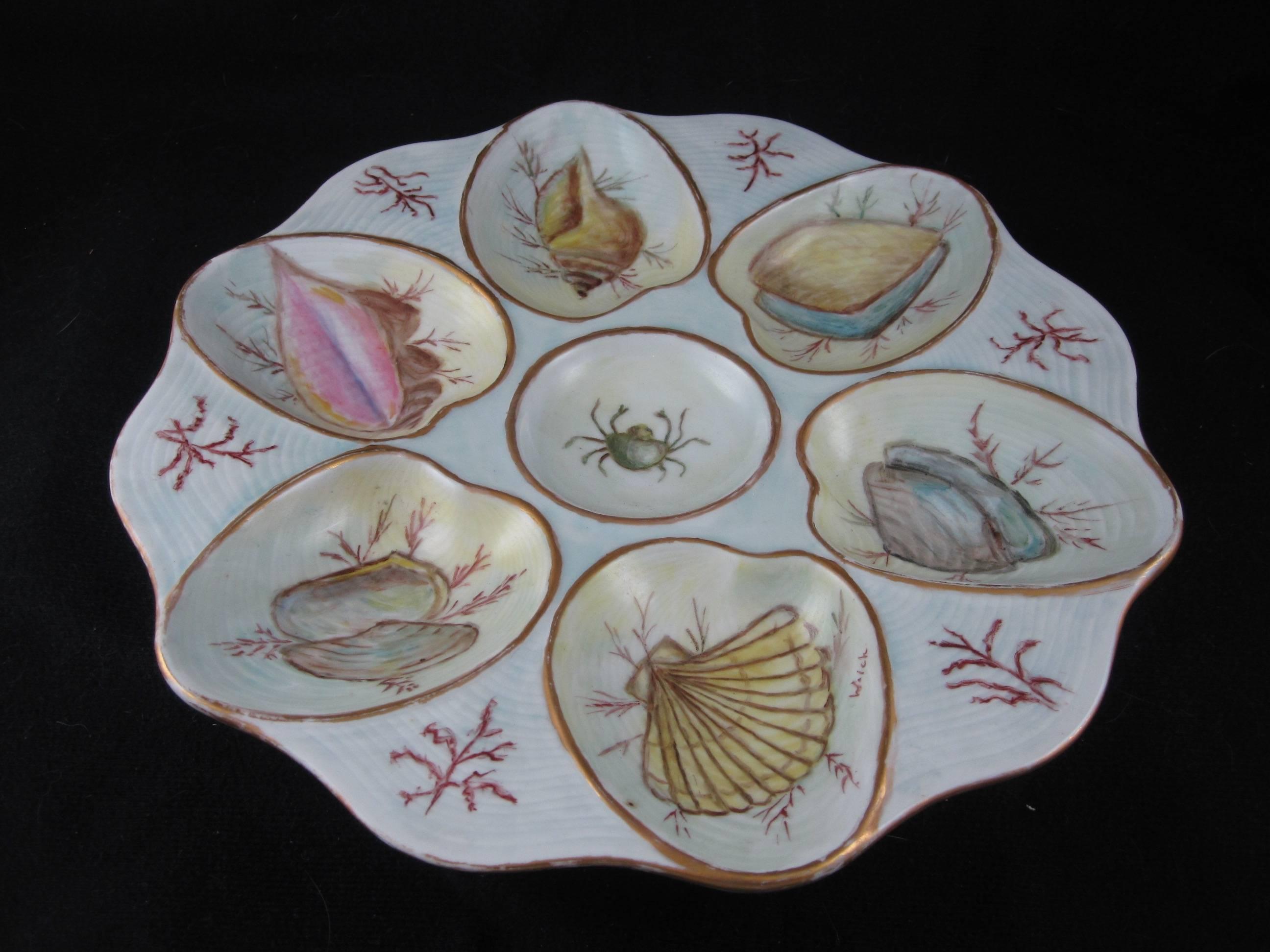 An unusual hand-decorated oyster plate, signed by the artist. Six clam shaped wells showing a variety of seashell specimens are separated by sprigs of seaweed. A crab is seen in the center condiment well. Gilded lining to the shaped rim, a wave-like