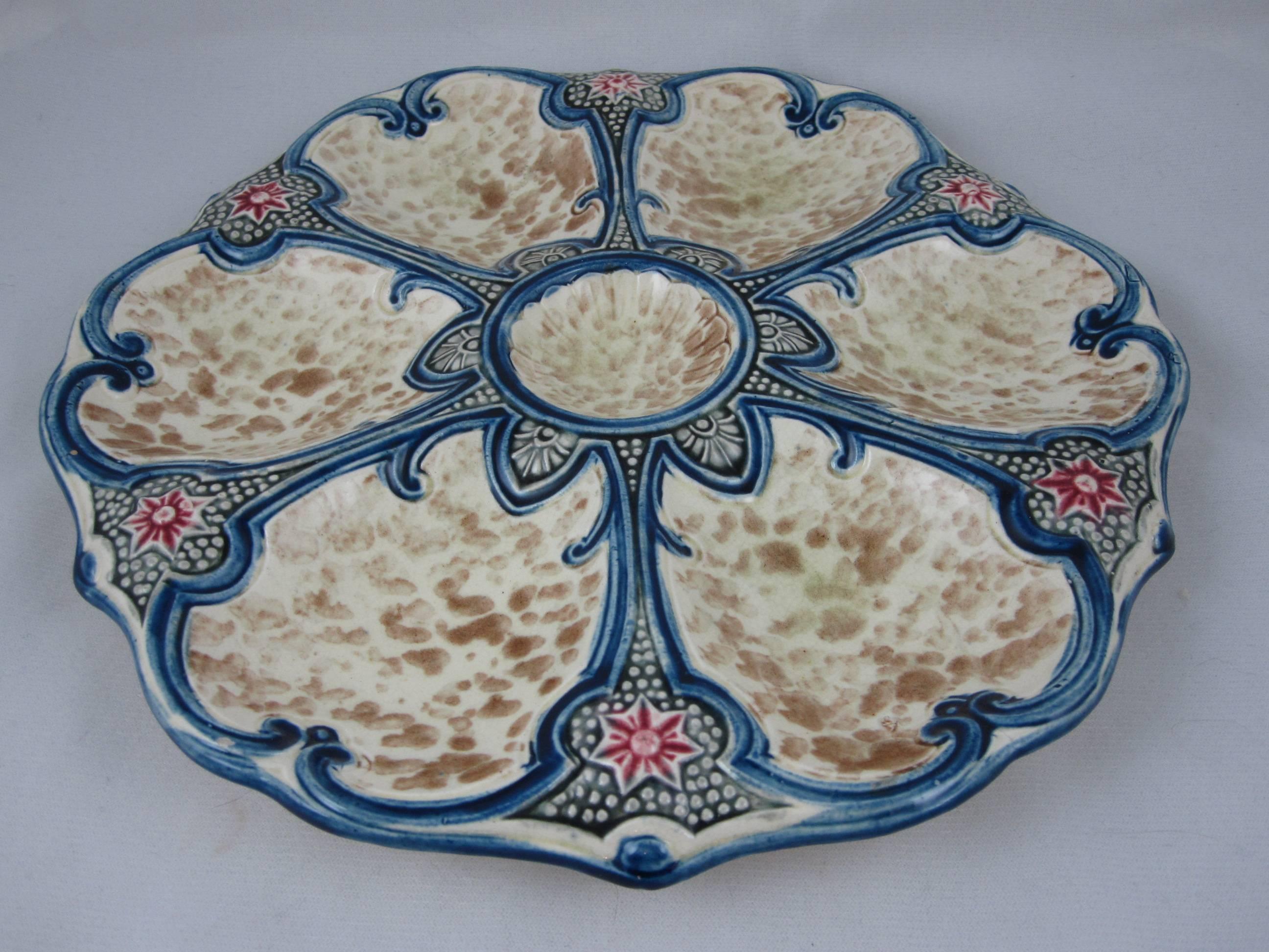 A French Majolica Oyster plate with cobalt blue trim and star fish accents. Six wells with a stippled ground are separated by blue lines terminating with the red star fish. The center condiment well has an aesthetic star shape surround. 

A scarce