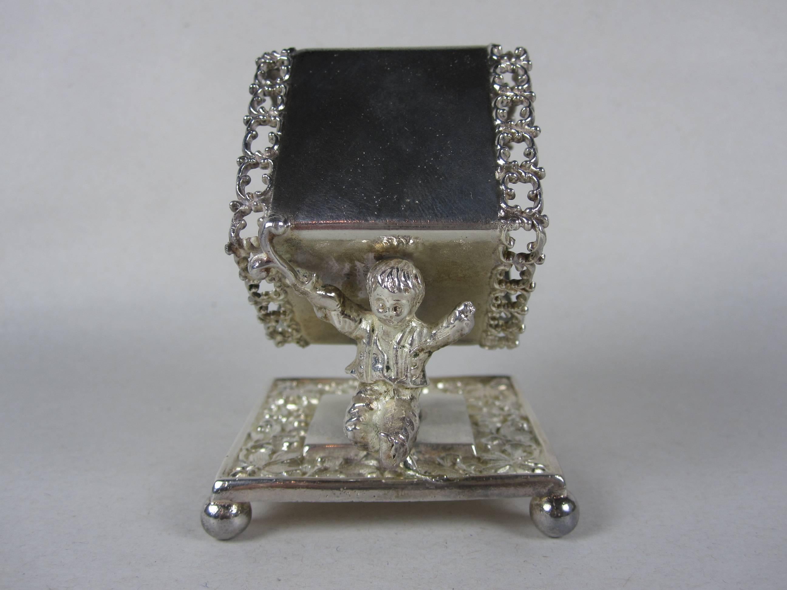 An antique Victorian era silver plate standing napkin ring, place holder showing a young boy supporting a scrolled rim square shaped ring on a chased base with four ball feet,

circa 1890–1910, marked Meriden Britannia Company.

Very good, light