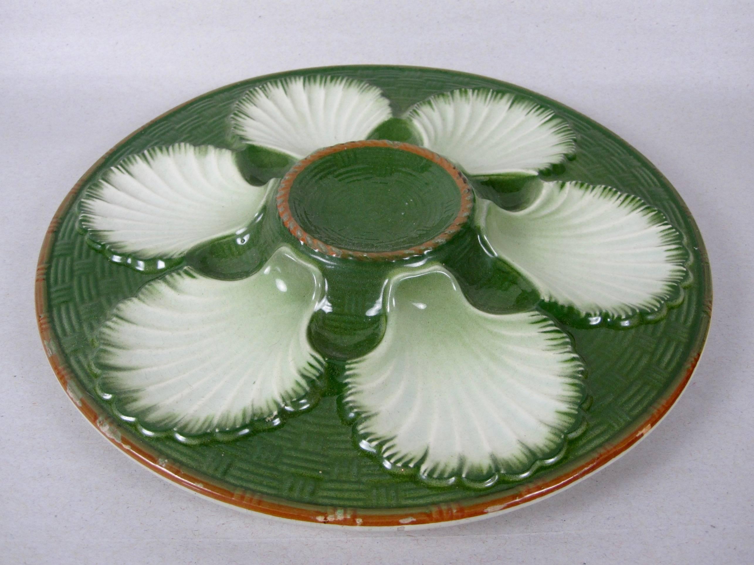 A French majolica glazed oyster plate with six scallop shell shaped wells on a deep forest green basket weave ground. A raised center condiment well, the outer rim of the center well and the plate showing the brown clay. 

Marked St. Clement, made