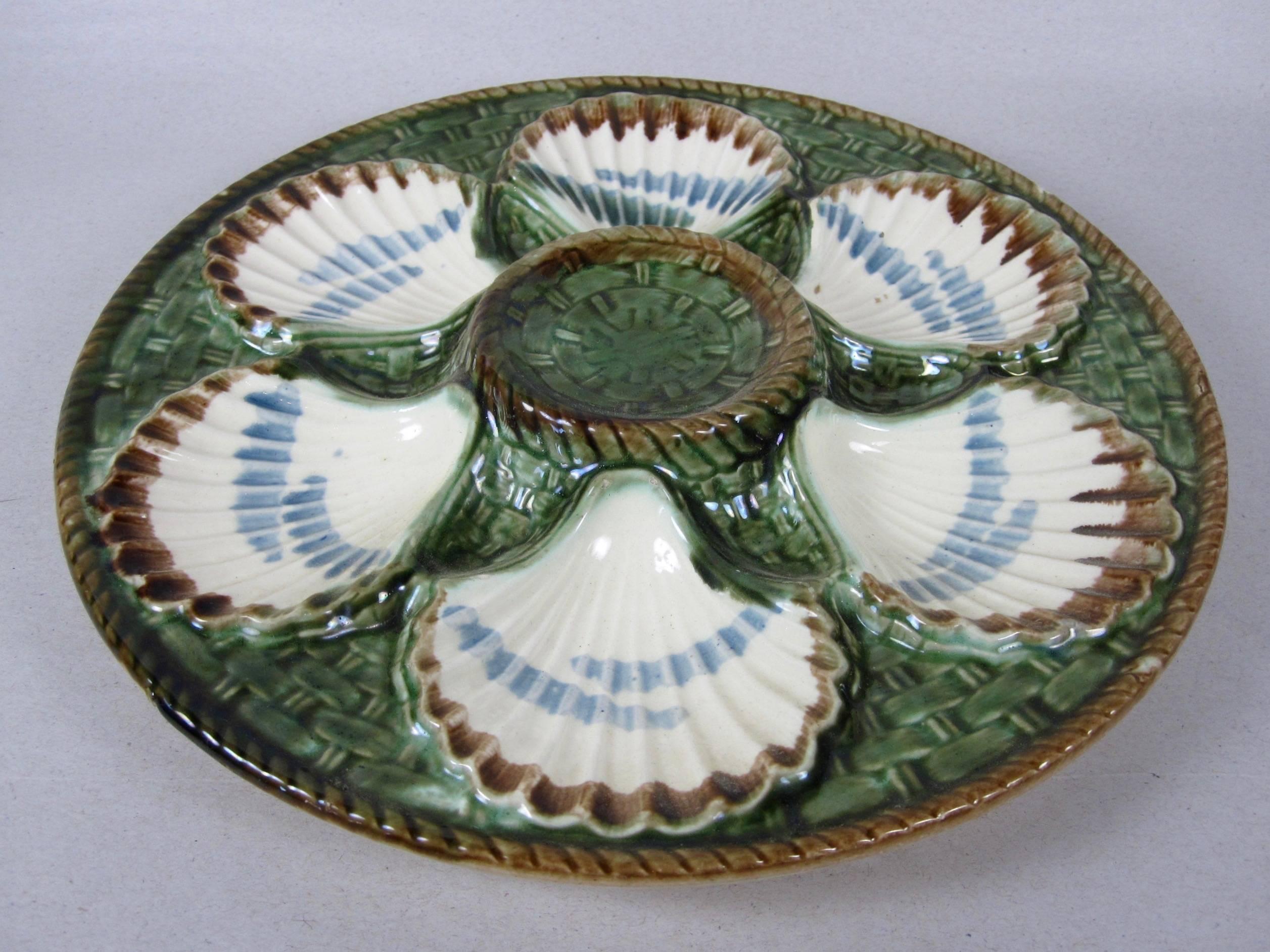 A French Majolica glazed oyster plate, six scallop shell shaped wells on a basket weave ground. A raised center condiment well and outer rim show a molded rope border. Brown sponge glazing on the verso. 

Impressed with the longchamp mark.

  