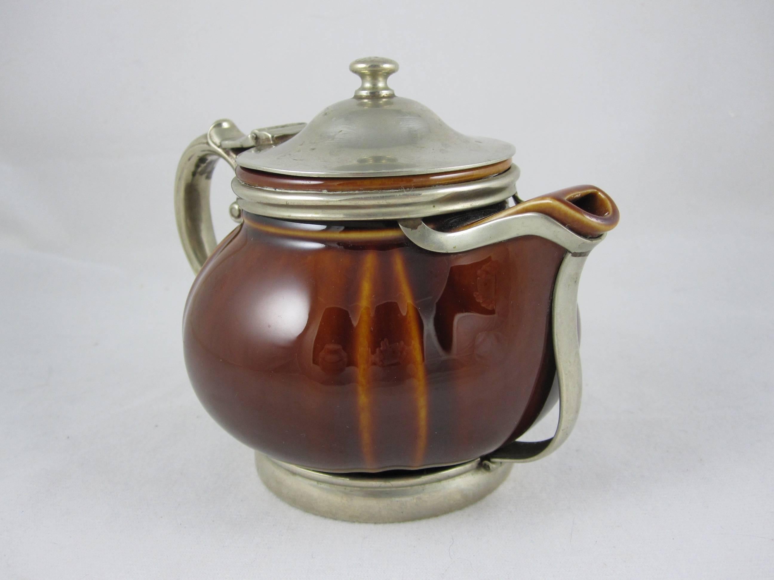 A sturdy hotel teapot, sized to hold one perfectly brewed cup of tea. The ceramic pot is braced in a silver soldered, silver plate holder of a base, handle and a lid with Art Deco styling. Glazed in a treacle-like brown, lighter on the raised mold