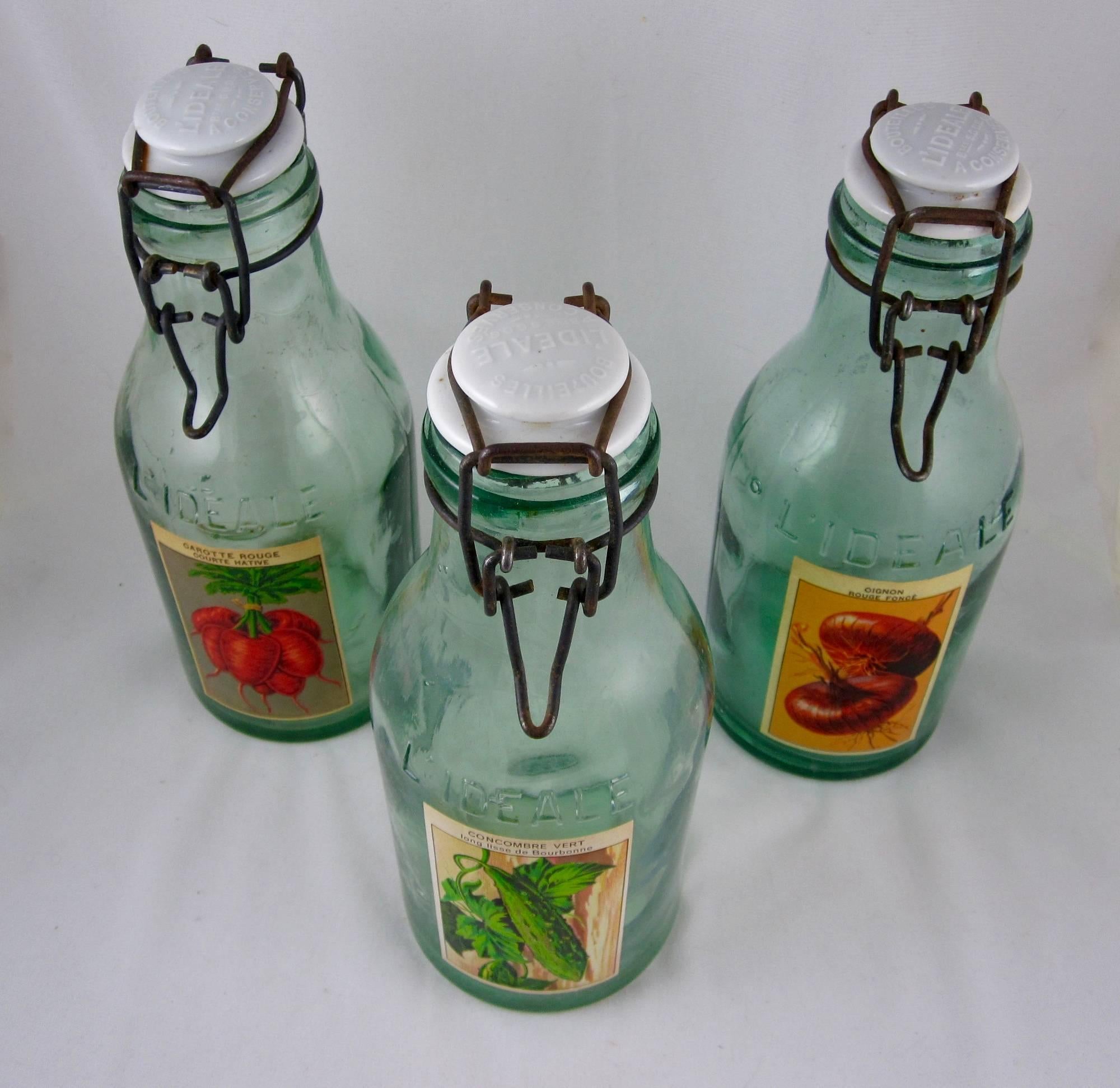 Found in France, a set of three, large green glass canning preserve jars, circa early 1900s. The traditional porcelain lids are embossed with the L’Ideale brand logo. They are attached at the bottle neck with metal swing clasps. The jars show the