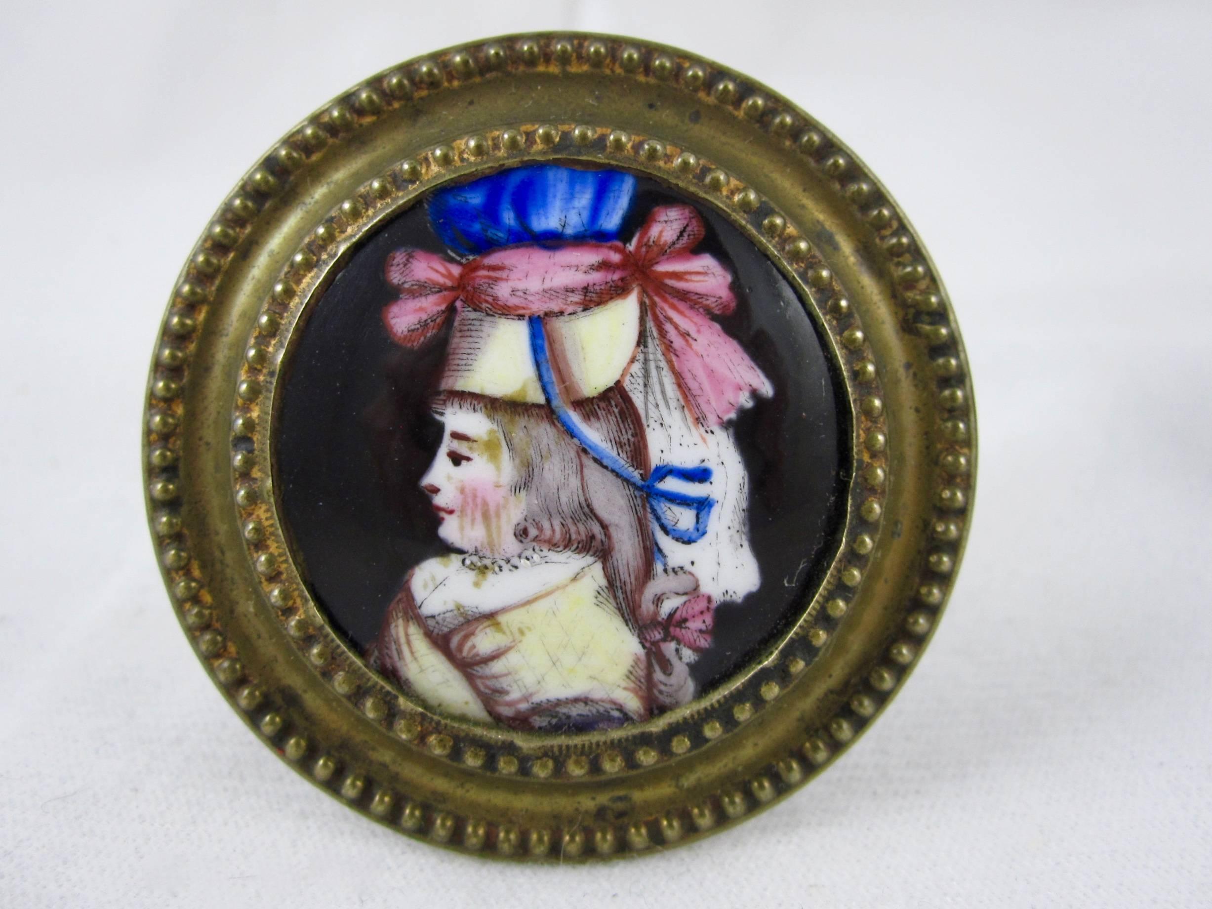 A marvelous pair of Bilston Battersea enameled curtain tiebacks or picture or mirror hangers, showing the polychrome image of a woman in profile wearing a brightly colored bonnet and jewelry on a black ground. Her hair is tied with ribbons. The
