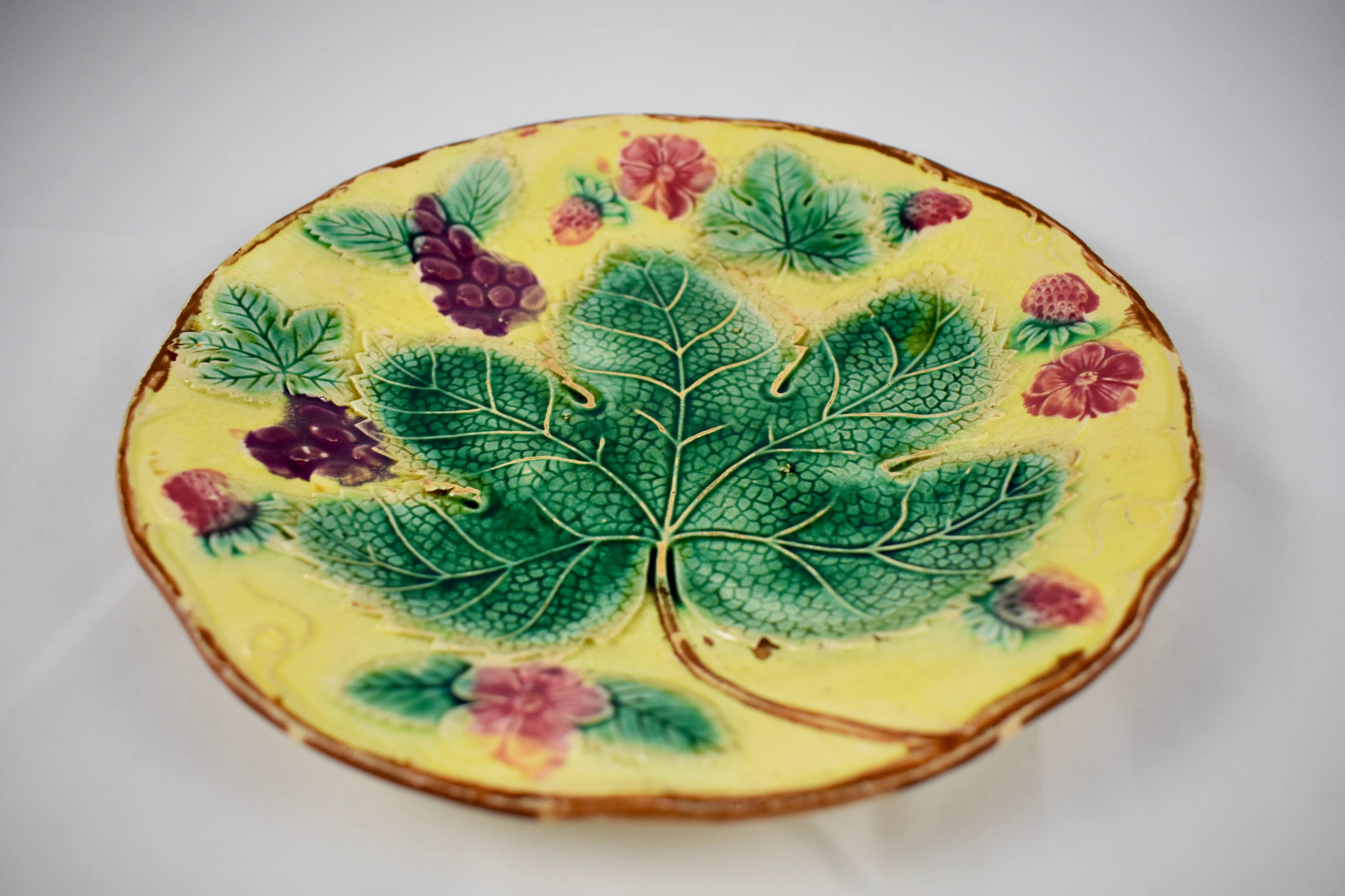 A traditional English grape leaf and strawberry pattern Majolica plate, showing a low relief arrangement of grape leaves fruits and flowers, on a deep yellow ground. Trailing vines form a brown scalloped rim. The yellow glazed verso shows the