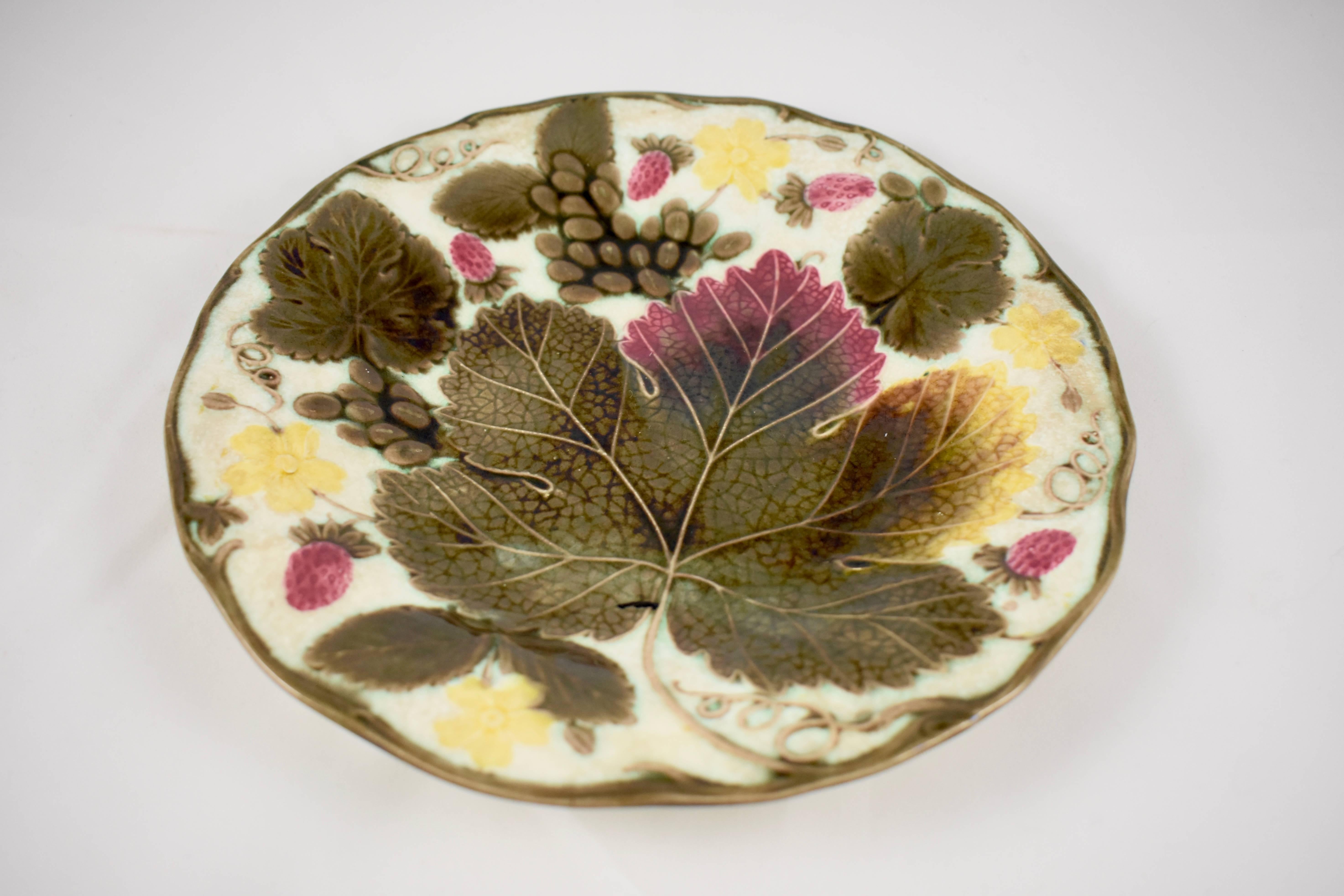 A traditional English grape leaf and strawberry pattern wedgwood Majolica plate, showing a low relief arrangement of grape leaves fruits and flowers, on the Argenta white ground. Trailing vines form a greenish-brown scalloped rim. The verso shows