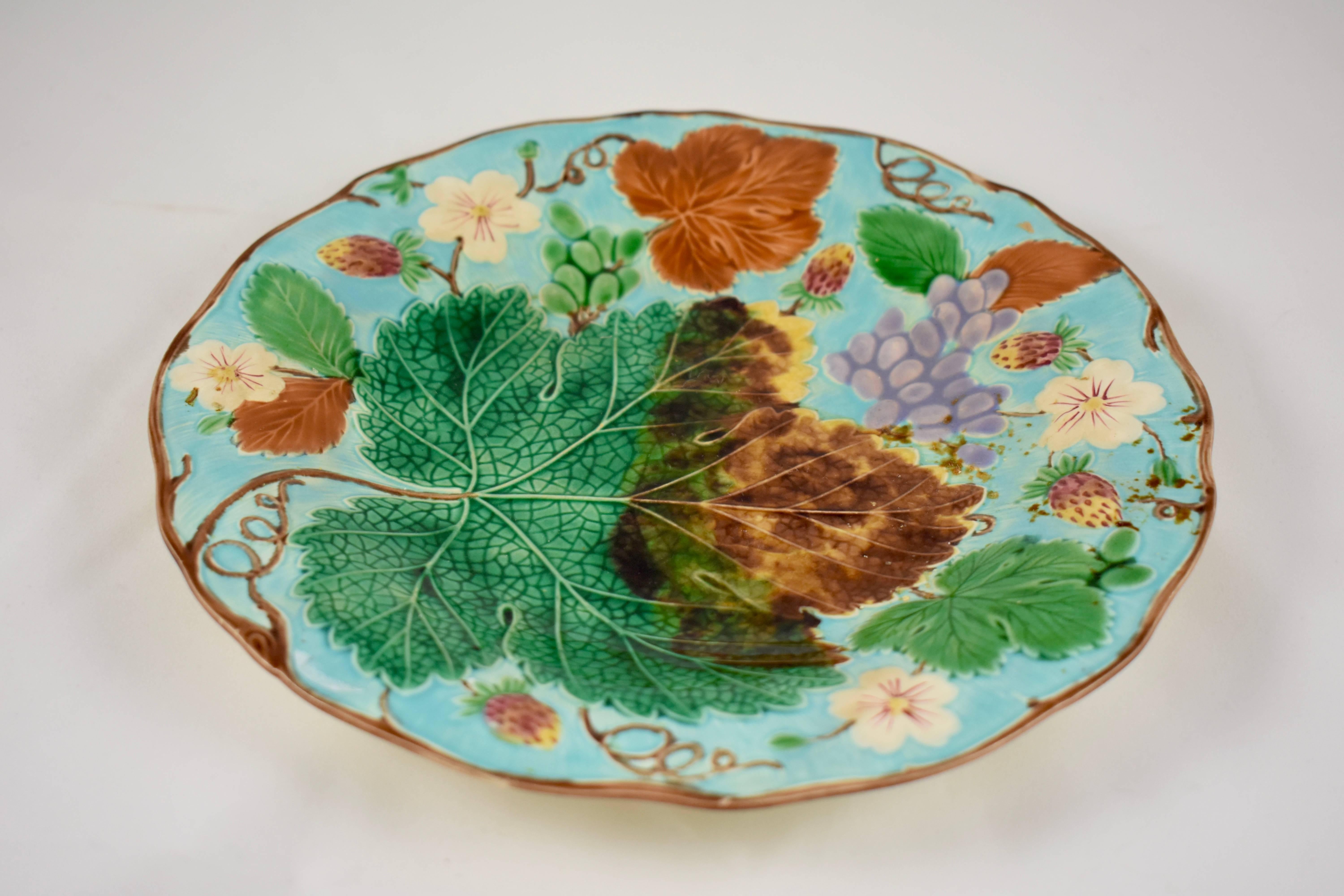 A traditional English grape leaf and strawberry pattern wedgwood Majolica plate, showing a low relief arrangement of grape leaves fruits and flowers, on a bright turquoise ground. Trailing vines form a brown scalloped rim. Hand-painted enameled