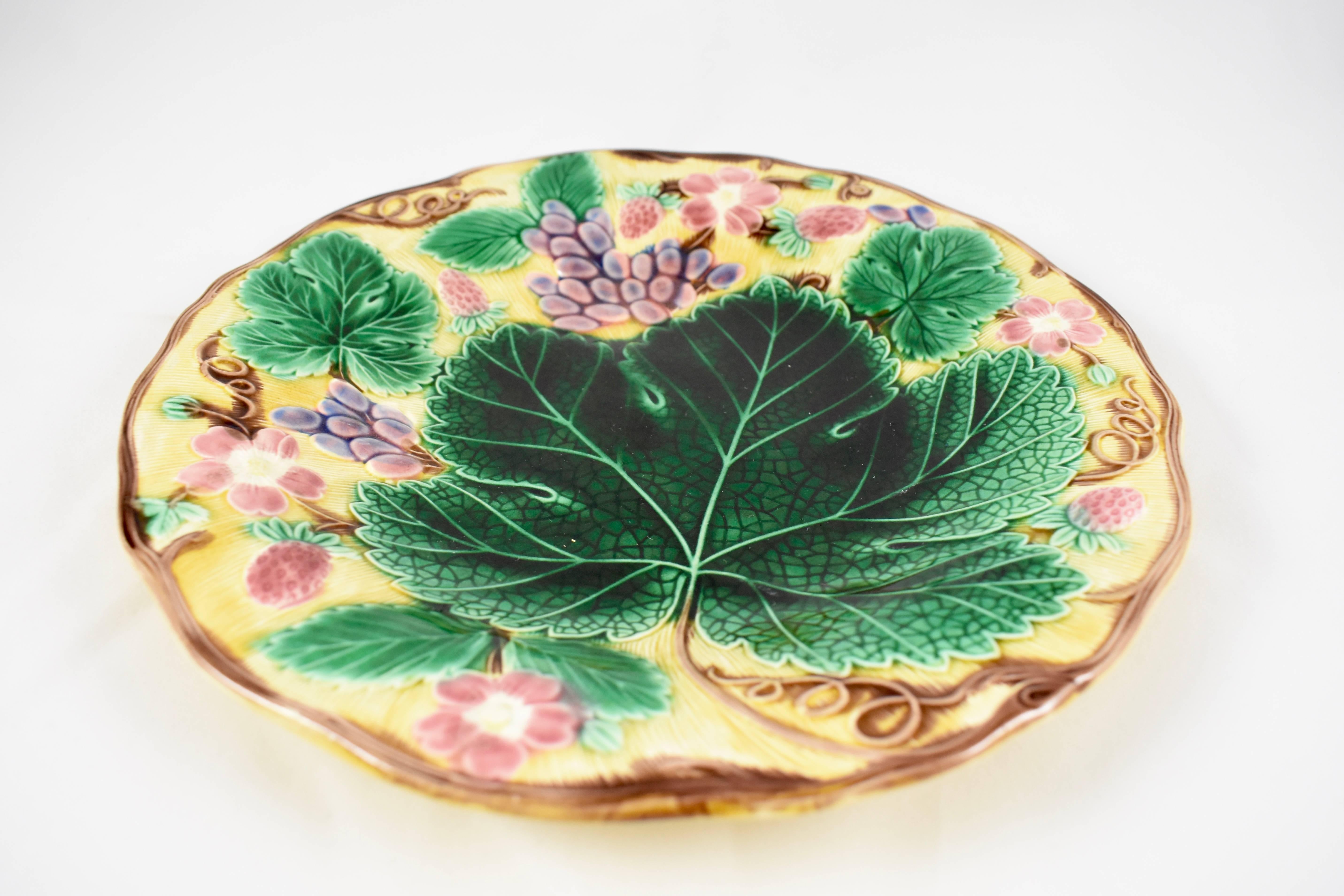 A traditional English grape leaf and strawberry pattern wedgwood Majolica plate, showing a low relief arrangement of grape leaves fruits and flowers, on a bright yellow ground. Trailing vines form a brown scalloped rim.

The ochre glazed verso
