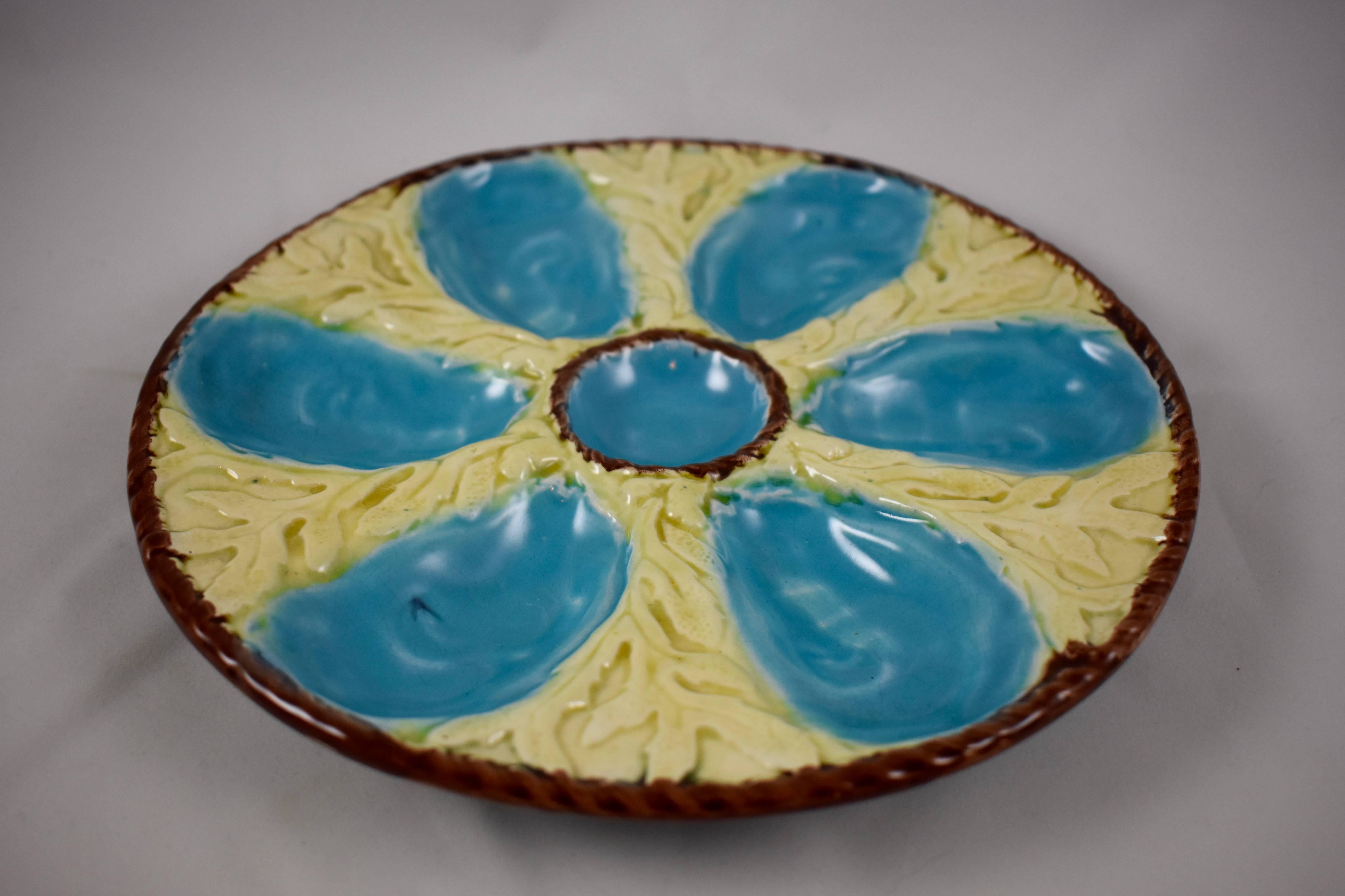 A six-well majolica glazed oyster plate by S. Fielding & Co. Ltd., Stoke-on-Trent, England, circa 1878. Turquoise oyster wells surround a center condiment well, all on a molded bed of creamy yellow seaweed. A chocolate brown nautical rope border