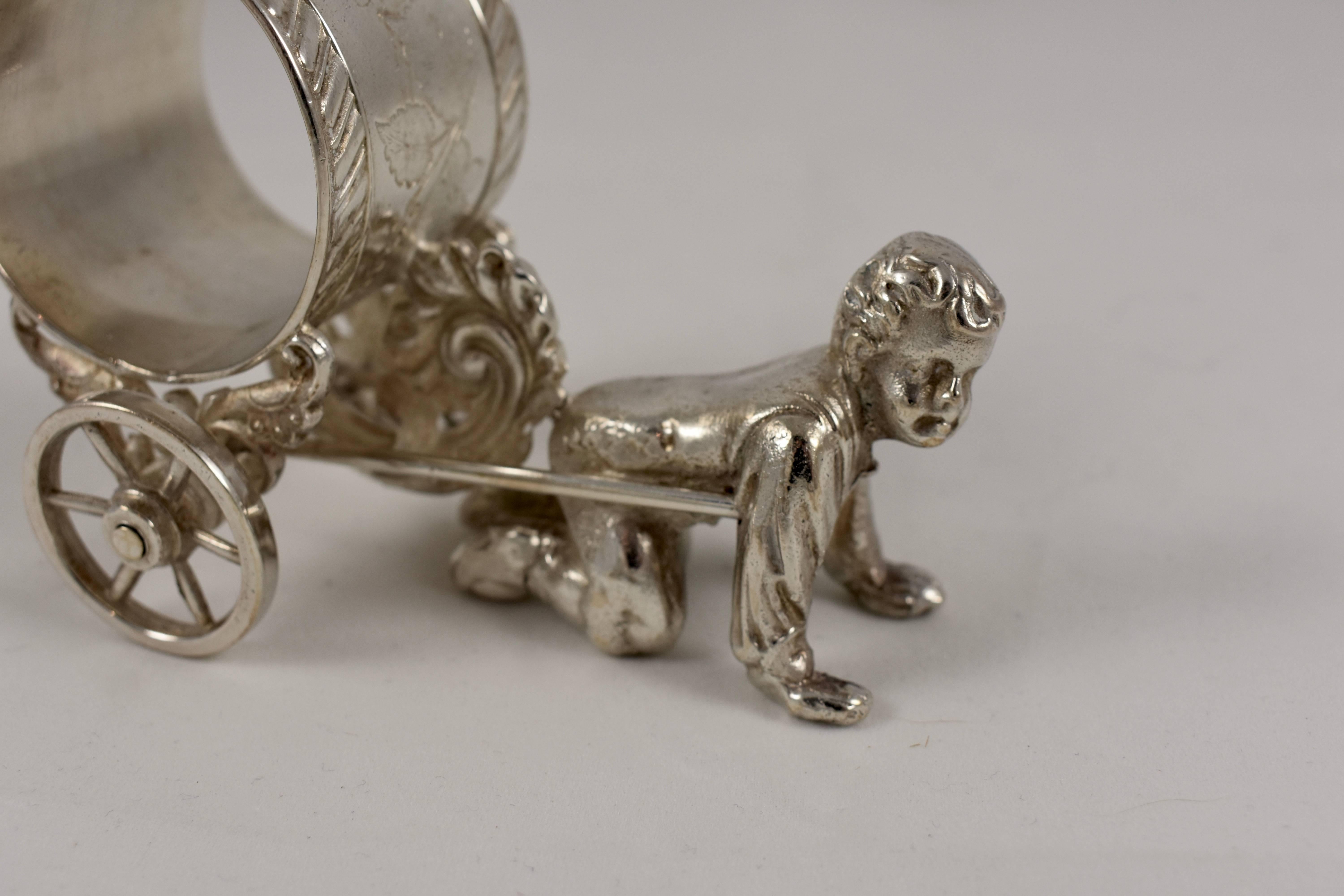 From the Victorian era, a silver plated figural napkin ring showing a young boy pulling a cart that holds the engraved ring. The child is dressed in knickers and a button-down shirt with a bow tied at his collar. The wheels on the cart are