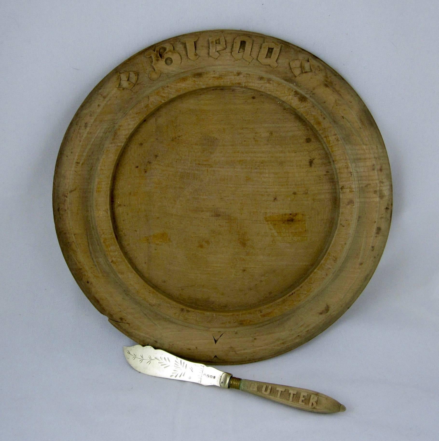A rustic 19th century round fruitwood bread board showing the word, 'Bread,' hand-carved into a stylized floral border with a deep inset rounded edge crumb catcher. Paired with a spreader, the wood handle carved with the word, 'Butter.' A floral