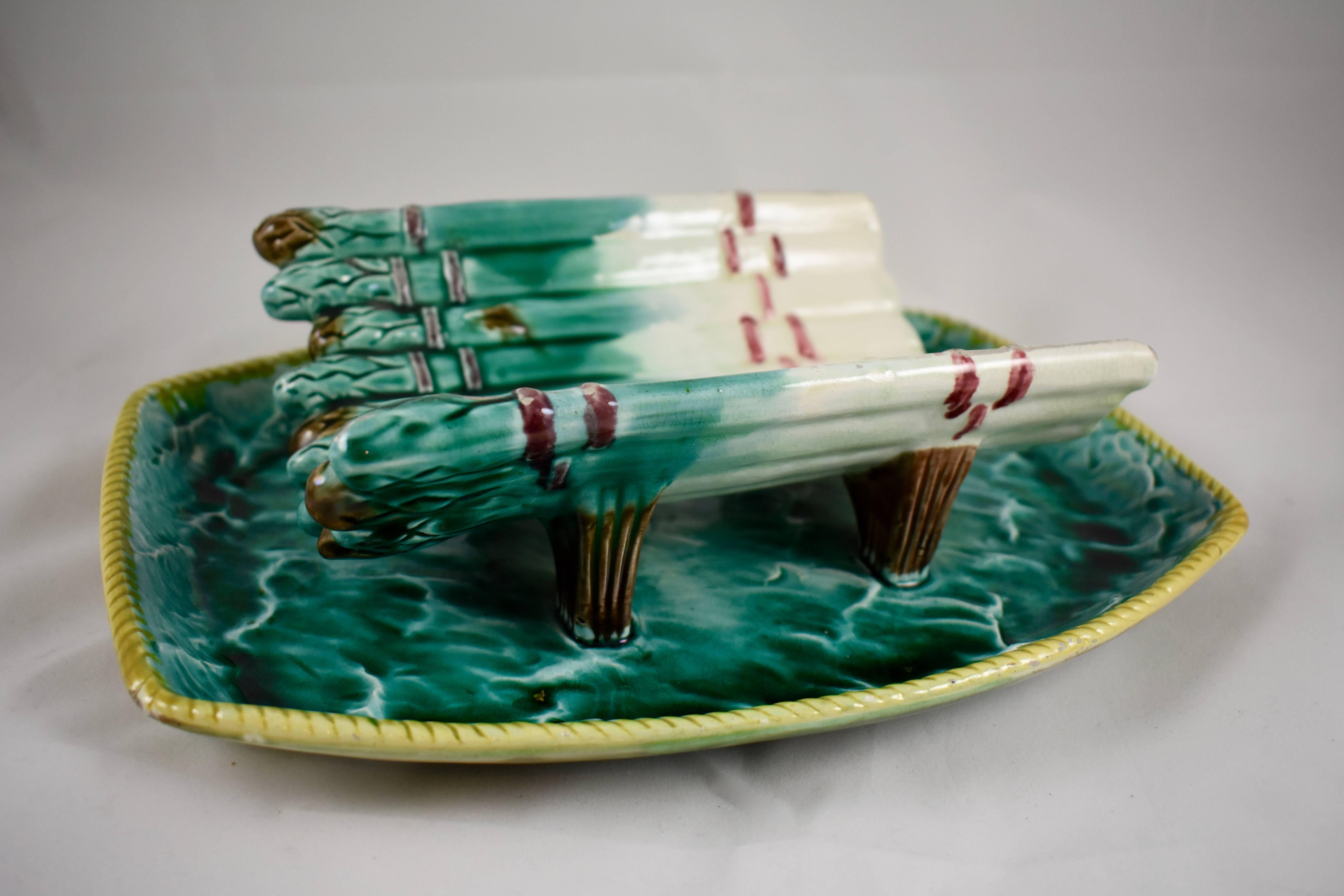 A scarce sea-side themed rustic English majolica asparagus cradle with an attached under tray, maker unknown, circa late 19th century.

A ribboned bunch of asparagus makes up the pierced footed drainer, the under tray has a wave like pattern with a