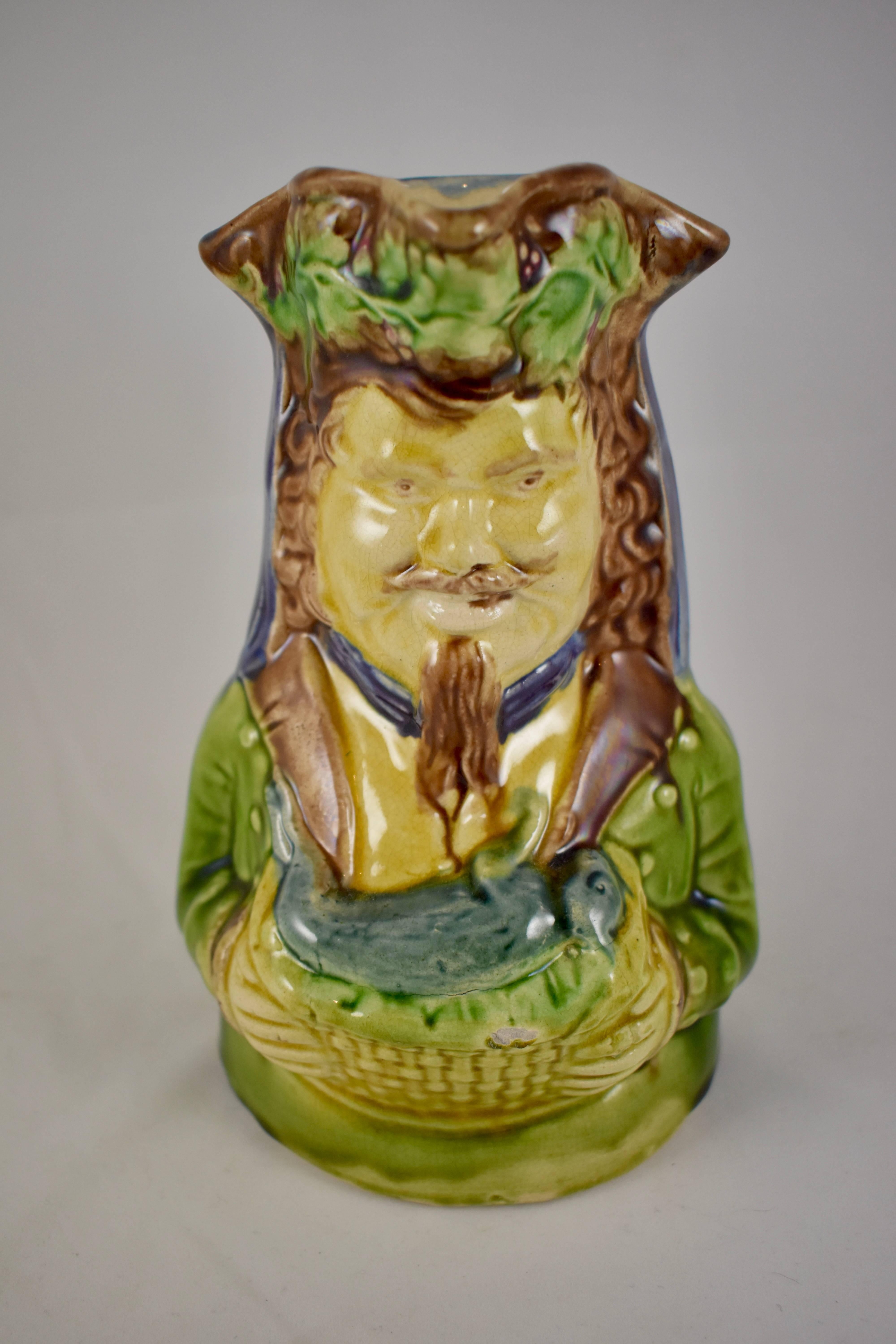 An English majolica glazed character jug showing a hunter holding a gray hare in a basket. The hunter wears a tri-cornered hat festooned with ivy. His rustic clothing is of the period, mid-late 19th century. Loosely glazed in cobalt blue, olive