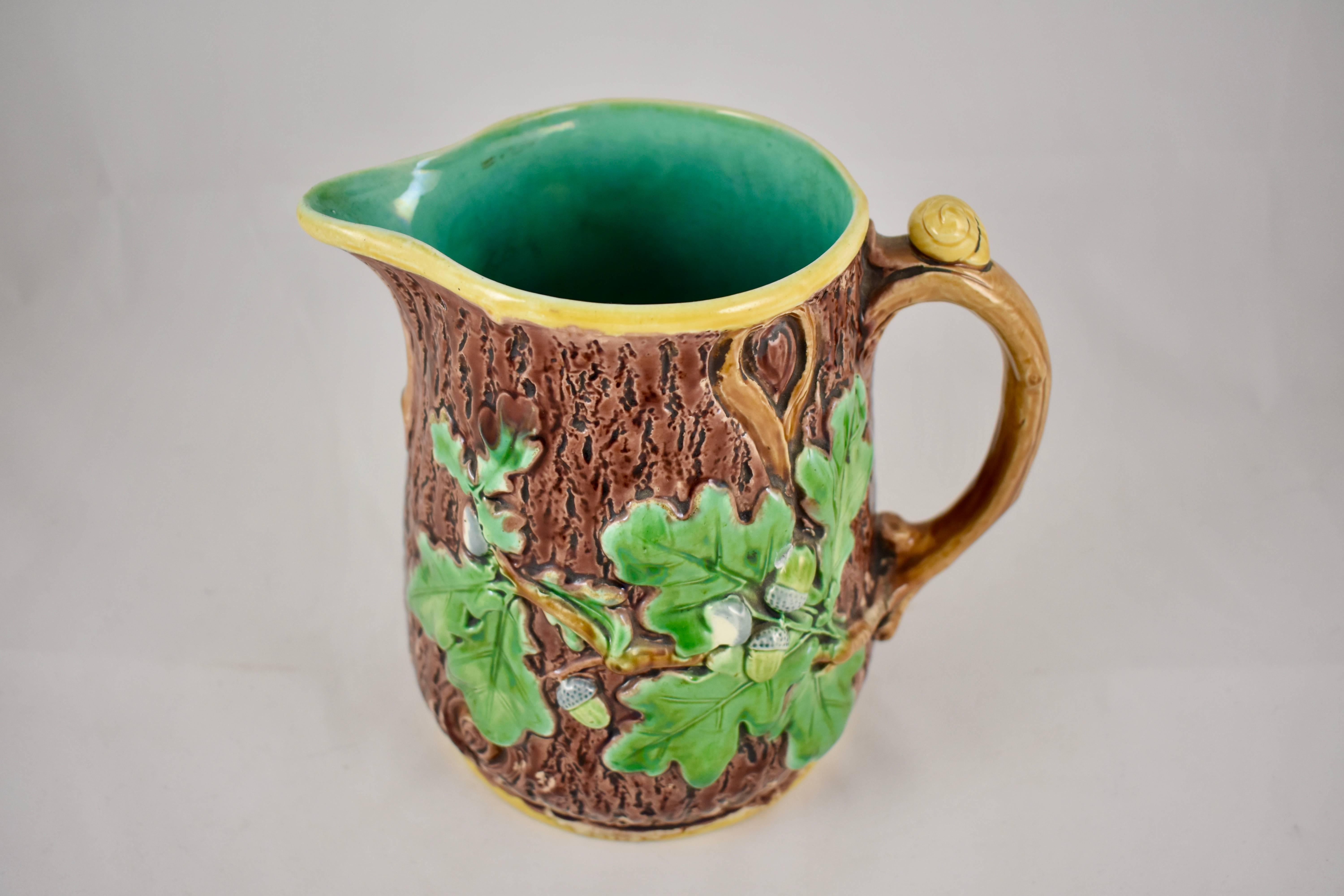 A Minton earthenware, majolica glazed oak leaf and snail pitcher, formed with a rustic brown tree trunk body embellished with oak leaves and acorns. The branch handle has a snail thumb set. The jug is rimmed with a yellow ochre band and has a