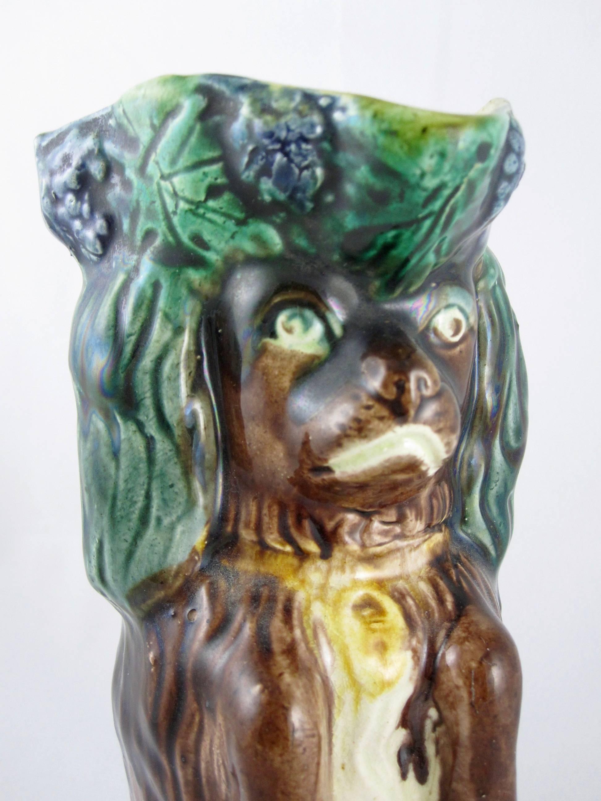 A Victorian, circa 1880 Majolica pitcher in the form of a begging King Charles Spaniel wearing a Tricorn hat decorated with bunches of grapes and their leaves. He also wears a padlock collar and the melting expression the Cavalier is known for. The