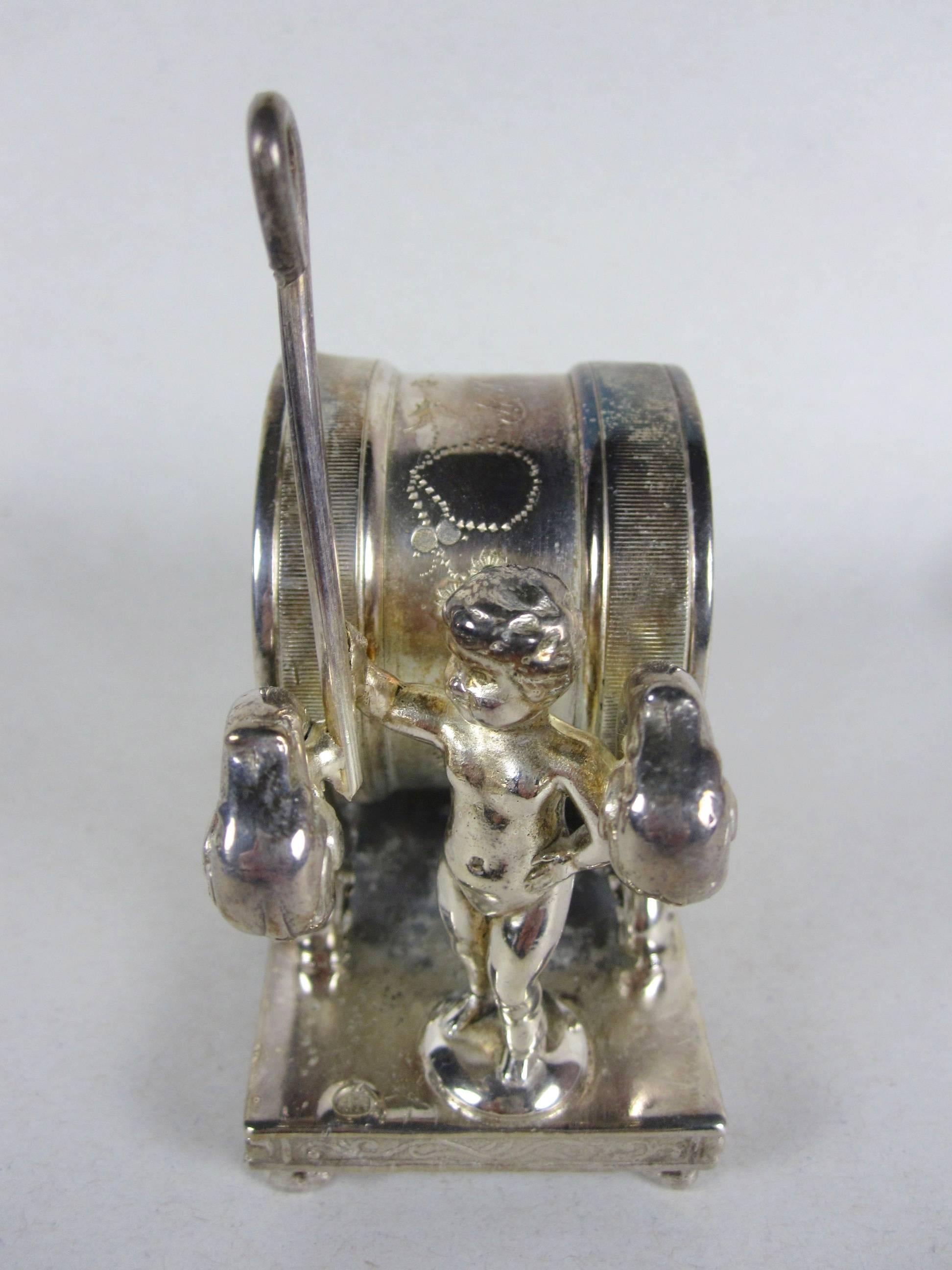 An antique Victorian era silver plate standing napkin ring place holder with a marching putto holding a staff, in front of an engraved barrel ring on a scrolled and ornate rimmed base. Initialed EBP. 

Dating from 1890-1910. Marked “Wilcox Silver
