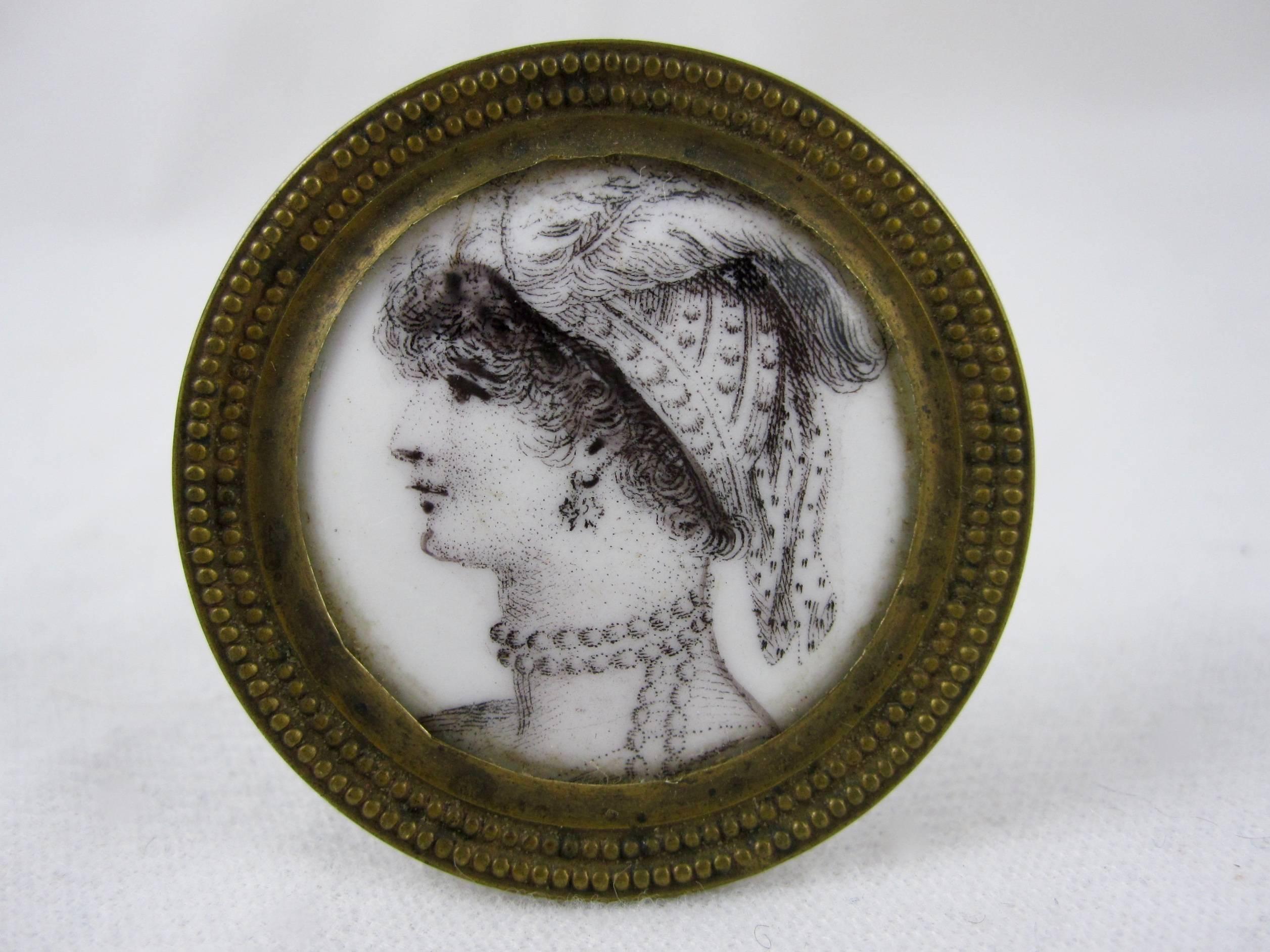 A marvelous pair of Bilston Battersea enameled curtain tiebacks, picture or mirror supports showing a transfer printed image of a woman in profile wearing a feathered head piece and jewelry. Grisaille coloring on a white ground set in a brass