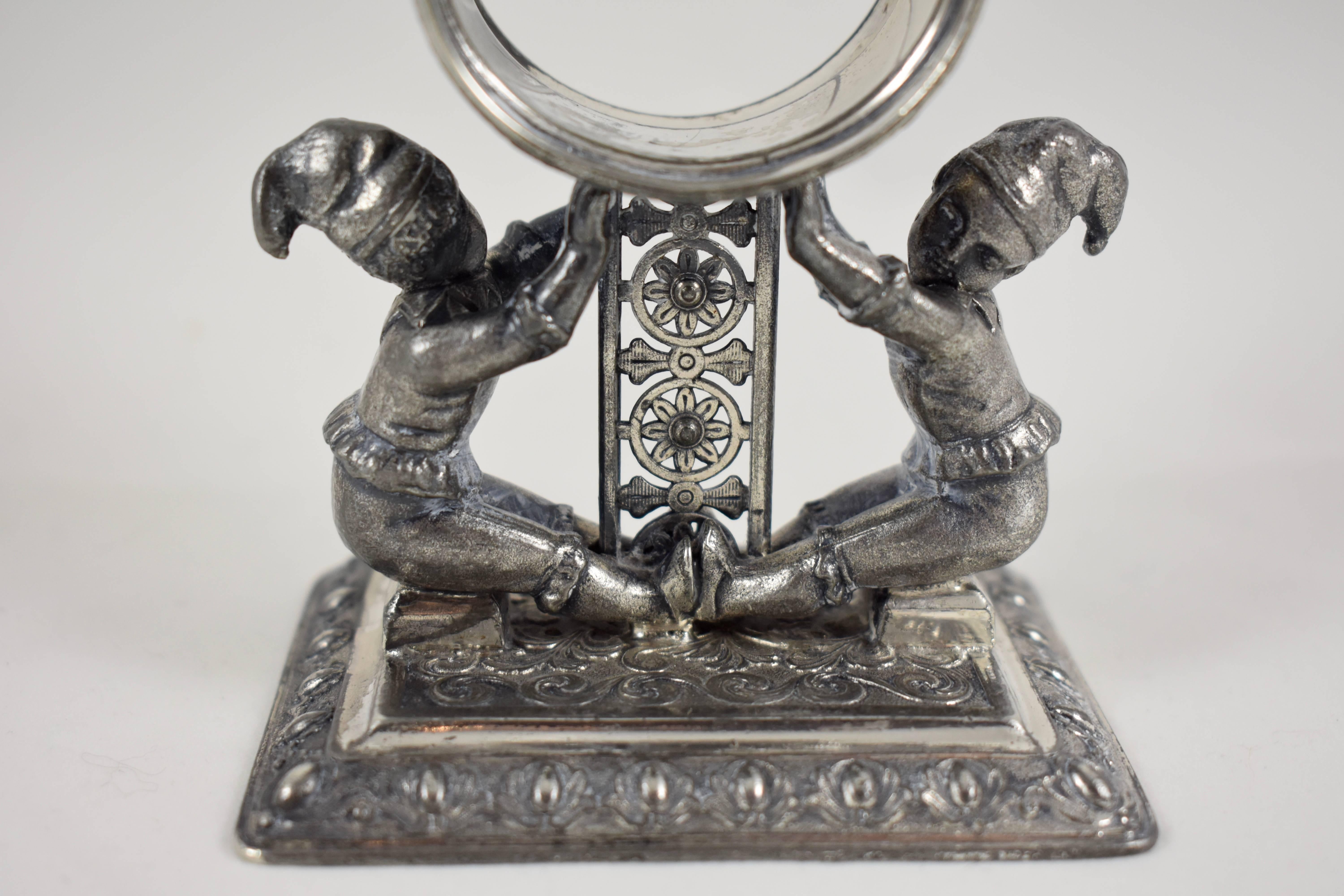 From the Victorian era, an Aesthetic movement silver plated figural napkin ring showing two jesters supporting a stylized floral column holding the heavily engraved ring. The base is ornately patterned as well. The figures, wearing stocking caps and