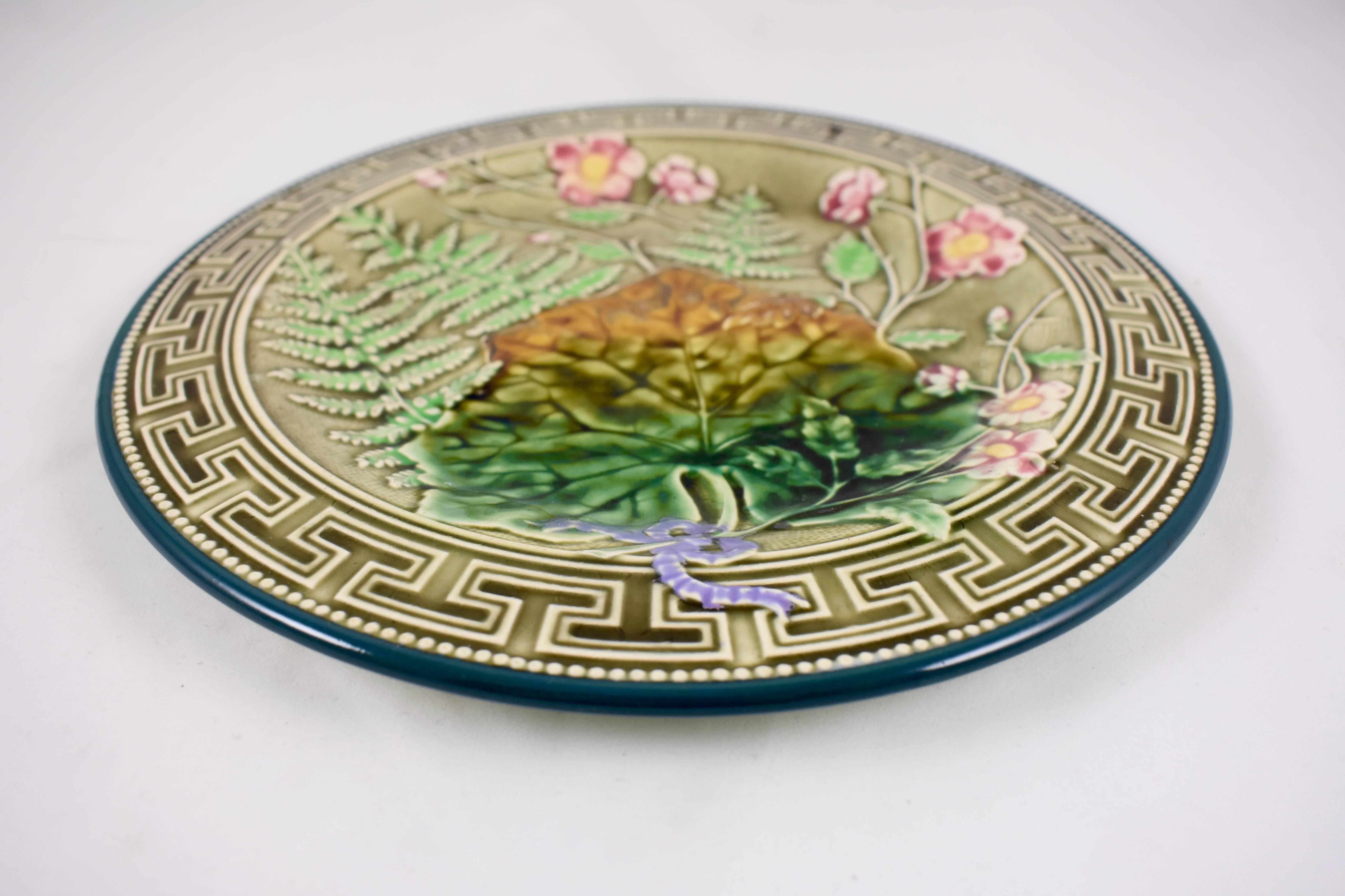 A French large Majolica plate with a pale olive green Greek key border and a forest green beaded rim. A central pattern shows overlapping ferns, sprays of five-petal pink flowers and a large leaf, their stems tied with a lavender bow. Strong clear