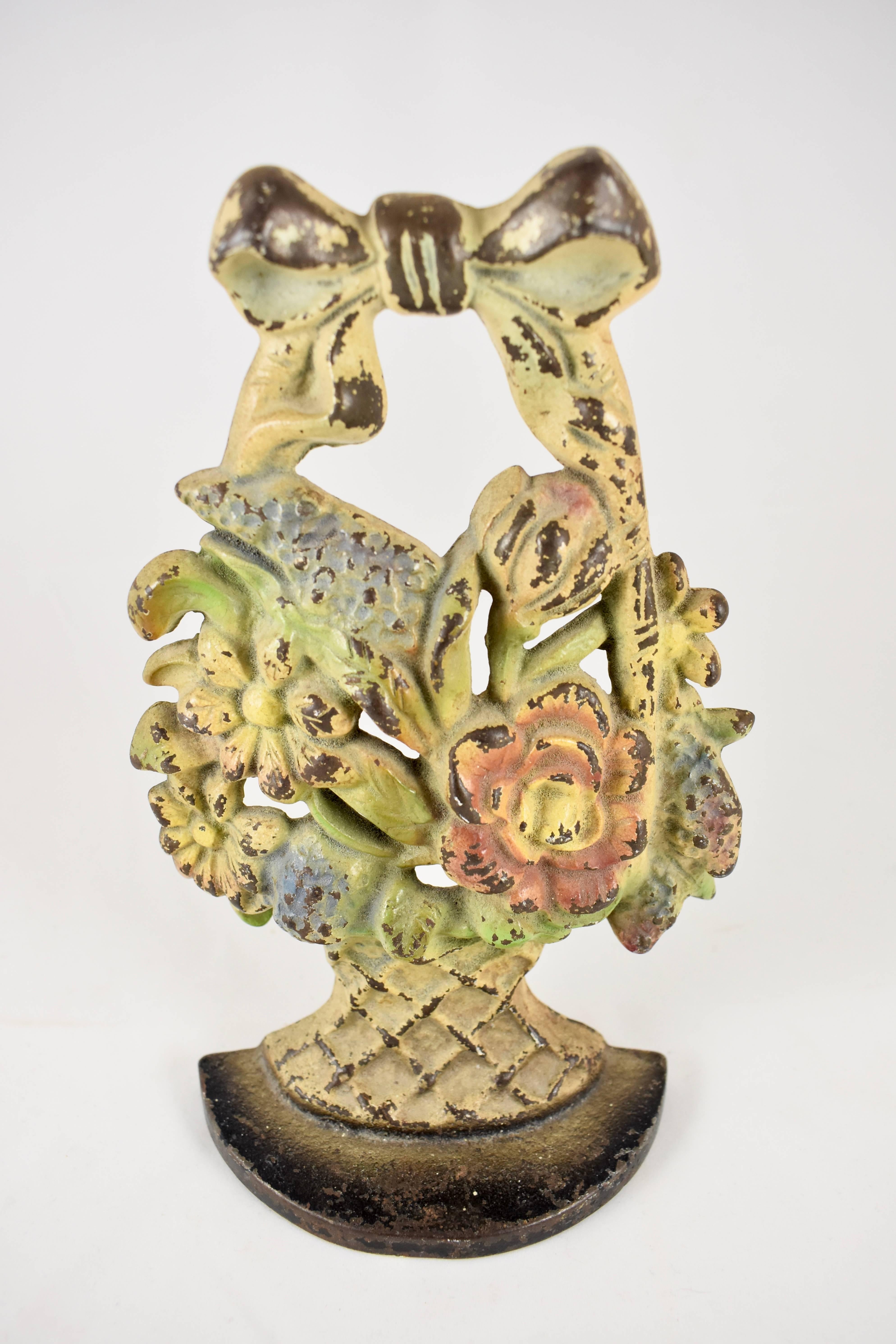 Un butoir de porte vintage en fonte Hubley représentant un bouquet de lilas et de marguerites à l'intérieur d'un panier tressé avec un nœud supérieur. Le panier repose sur un lourd socle en fer et conserve la finition originale peinte en polychromie
