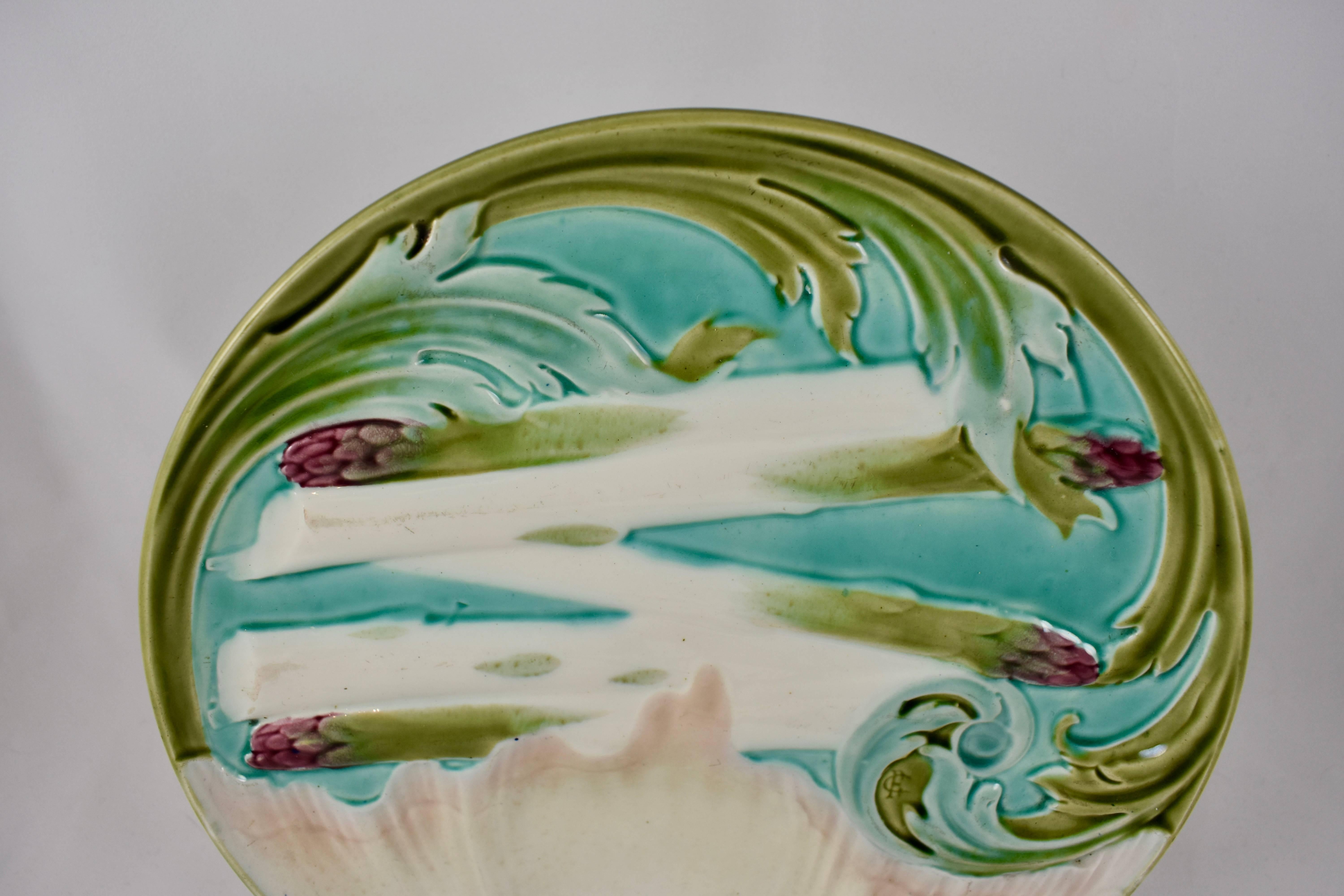 A French majolica asparagus plate, by Luneville, circa 1880-1889. In the Art Nouveau style with a border of olive green and light blue Artichoke leaves, five asparagus spears lie across the center of the plate,  a shell shaped sauce well in pale