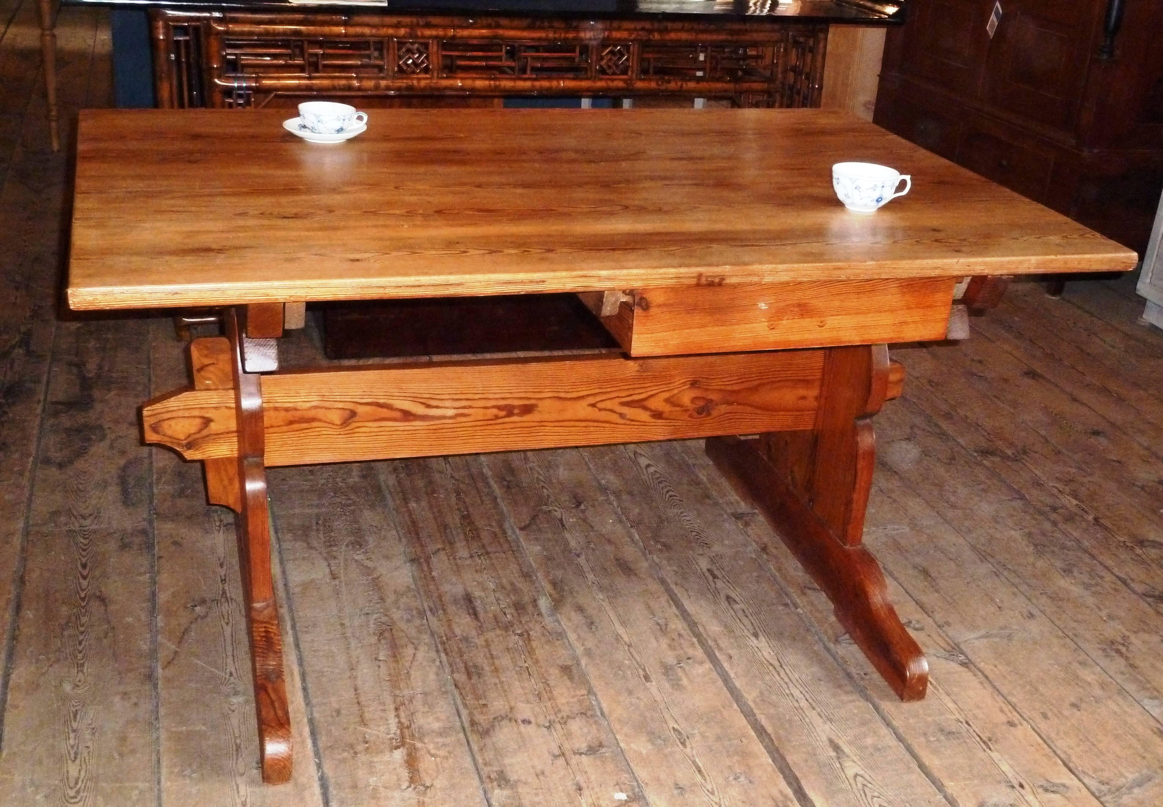 This antique Swedish country farmhouse table displays an unusual smaller size and a very charming handcrafted trestle base form. The table shows an expansive pine top with a beautifully grained warm patina and two large hanging dovetailed drawers